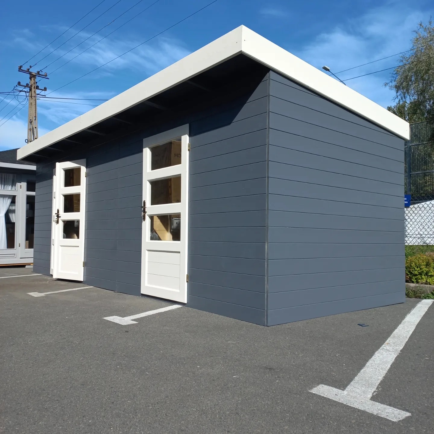 Nowoczesna, szara konstrukcja z dwoma białymi drzwiami z przeszkleniami, ustawiona na parkingu z widocznymi liniami, pod błękitnym niebem.