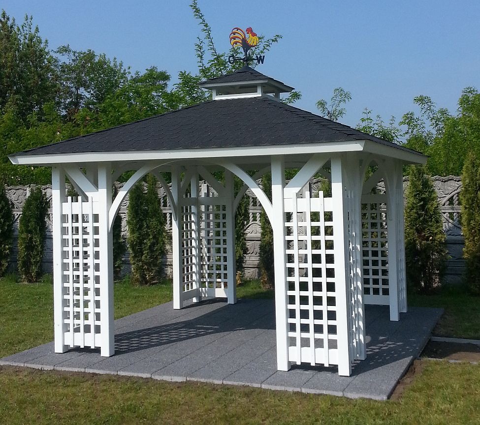 Biała altana ogrodowa z czarnym dachem, zwieńczona wiatrowskazem z kogutem, na szarym podłożu z kostki brukowej, otoczona zielenią.