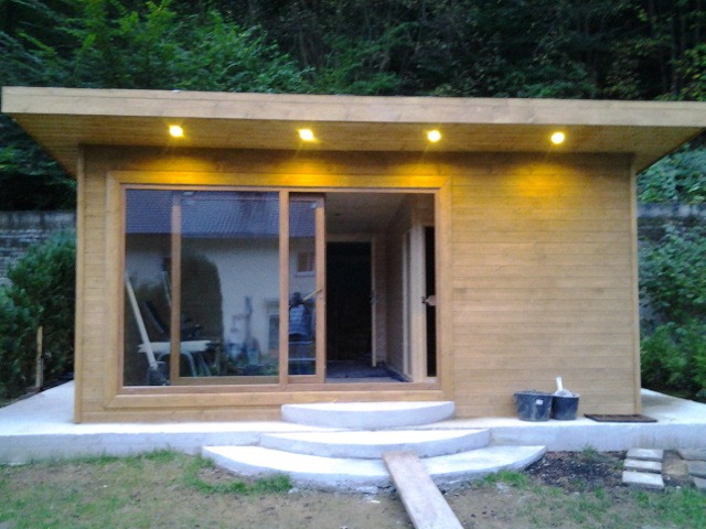 Nowoczesna, drewniana konstrukcja z płaskim dachem i dużymi przesuwnymi oknami, oświetlona punktowymi lampami, z betonowymi schodami wejściowymi i widocznymi narzędziami budowlanymi obok, otoczona...