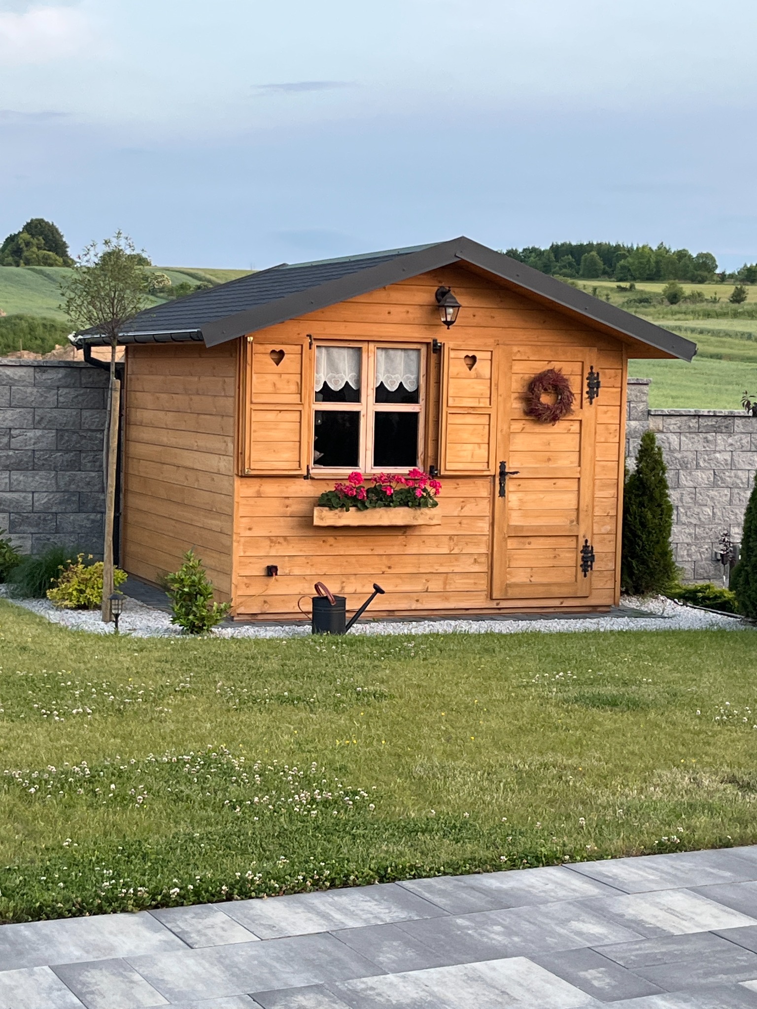 Drewniana konstrukcja z oknem i drzwiami, ozdobiona serduszkami i wiankiem, otoczona zielonym trawnikiem i kamiennym murem.