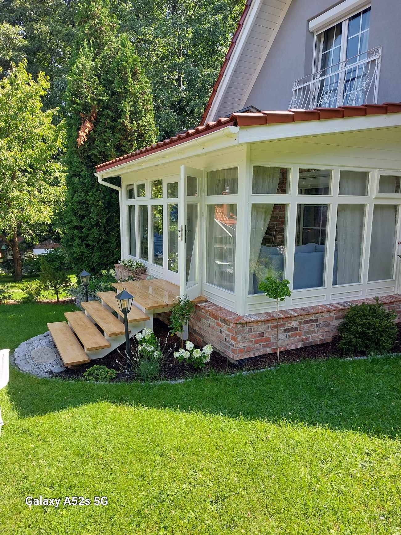 Zadaszony taras z drewnianymi schodami, ceglaną podstawą i białymi ramami okiennymi, otoczony zielenią i lampami ogrodowymi.