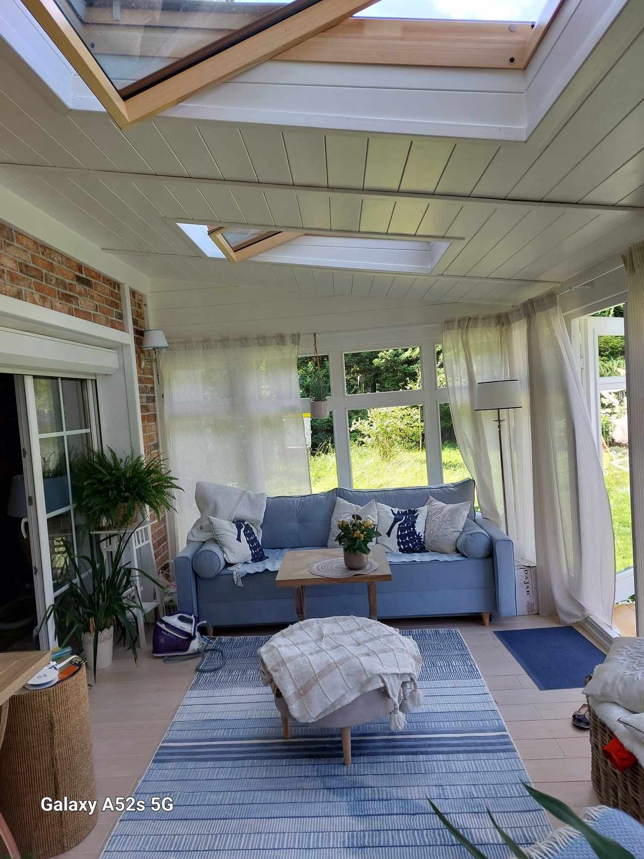 Jasne wnętrze z ceglanym akcentem, przeszklonym dachem i niebieską sofą z poduszkami w marynistycznym stylu. Widoczne rośliny doniczkowe i dywan w paski.