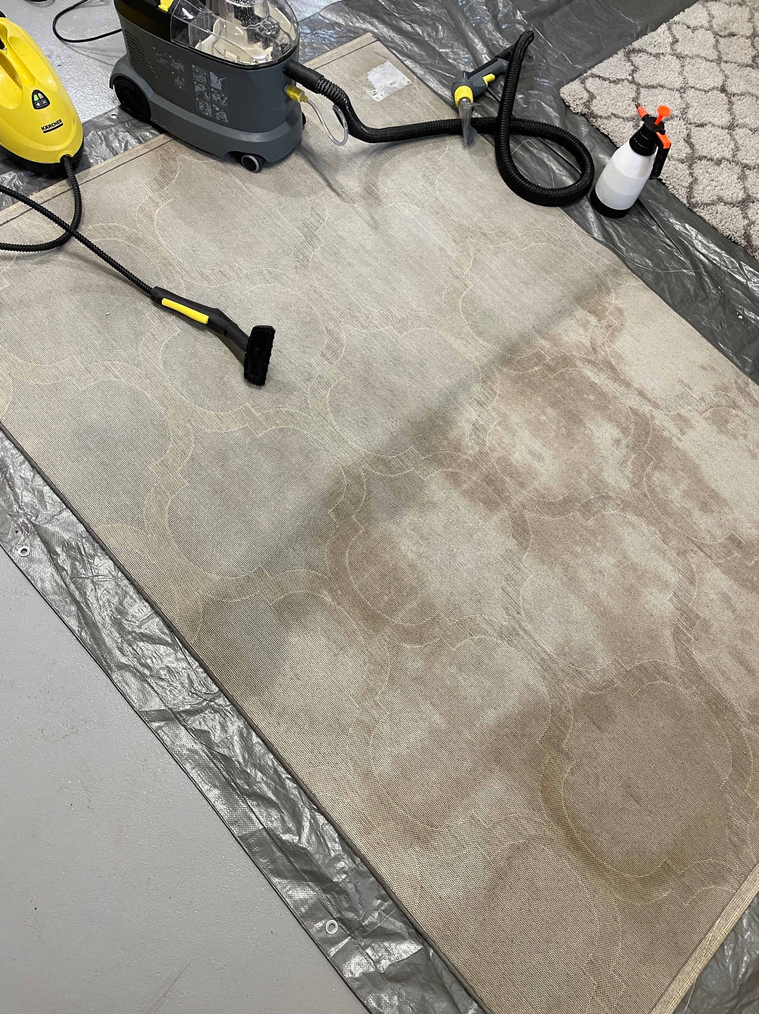 Beżowy dywan z wzorem w trakcie czyszczenia, widoczne plamy i urządzenia czyszczące na folii ochronnej.