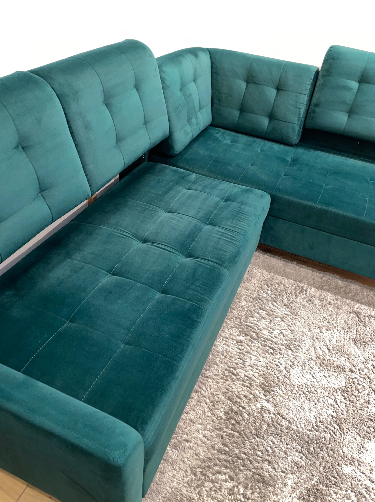 Fragment narożnika z zielonego weluru, z pikowanymi siedziskami i oparciami, ustawionego na puszystym, jasnym dywanie.