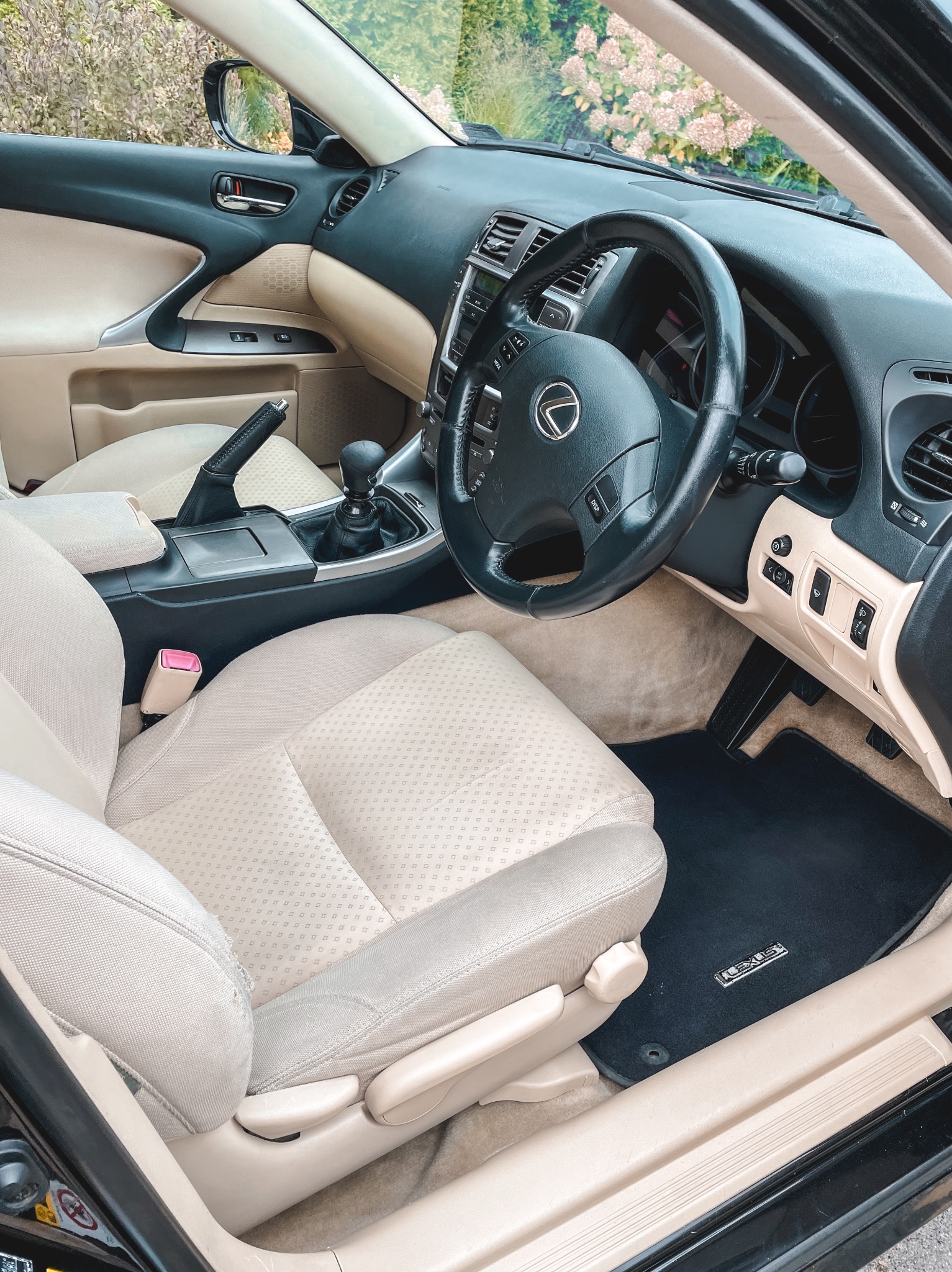 Jasne wnętrze samochodu Lexus z widocznymi beżowymi skórzanymi siedzeniami, czarną kierownicą z logo, czarnymi dywanikami i elementami deski rozdzielczej w kolorze beżowym i czarnym.