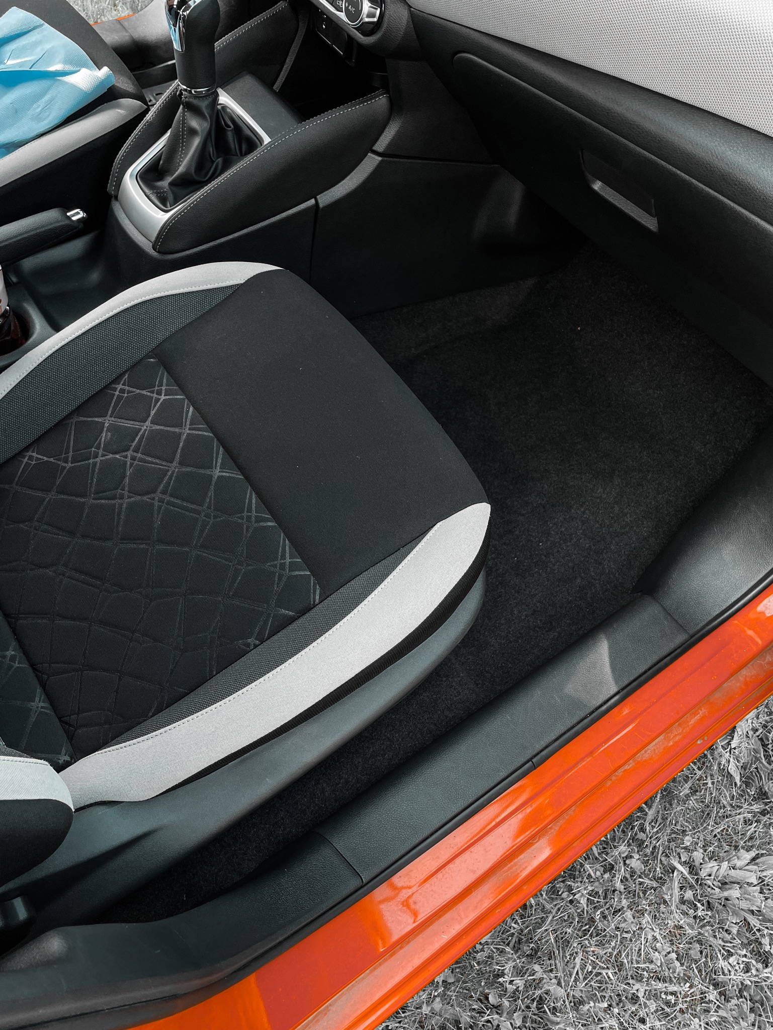 Wnętrze samochodu z czarną tapicerką fotela z geometrycznym wzorem i czarną wykładziną podłogową, widoczne elementy deski rozdzielczej i dźwigni zmiany biegów.