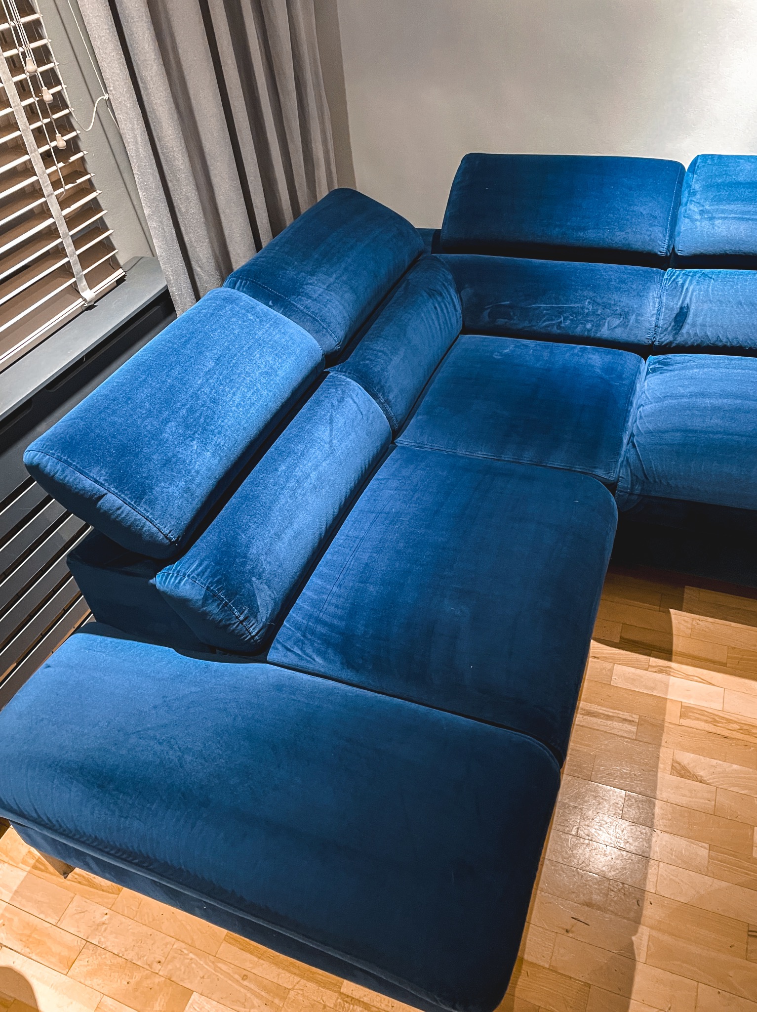 Narożnik modułowy w odcieniach niebieskiego aksamitu, ustawiony na parkiecie w pomieszczeniu z żaluzjami pionowymi i zasłoną.