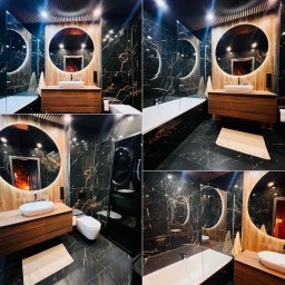 Elegancka łazienka z czarnymi płytkami imitującymi marmur, okrągłym lustrem z podświetleniem LED i drewnianą szafką pod umywalką. Nowoczesny design z akcentami drewna.