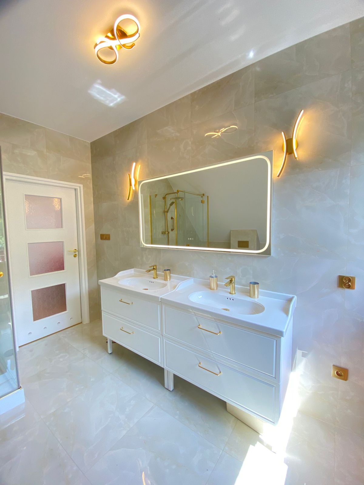 Jasna łazienka z dwoma umywalkami na białej szafce, lustrem z podświetleniem LED i złotymi akcentami. Widoczne płytki imitujące marmur i nowoczesne oświetlenie.