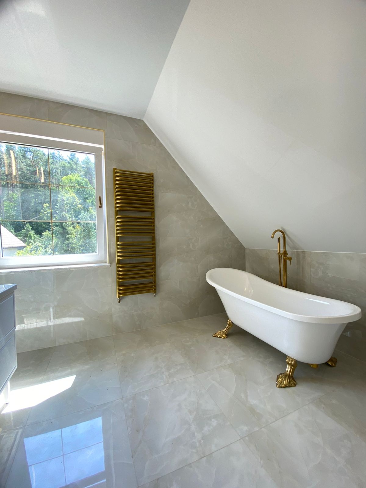 Jasna łazienka na poddaszu z wolnostojącą wanną na złotych nóżkach, złotym grzejnikiem i armaturą. Okno z widokiem na zieleń. Płytki imitujące marmur.