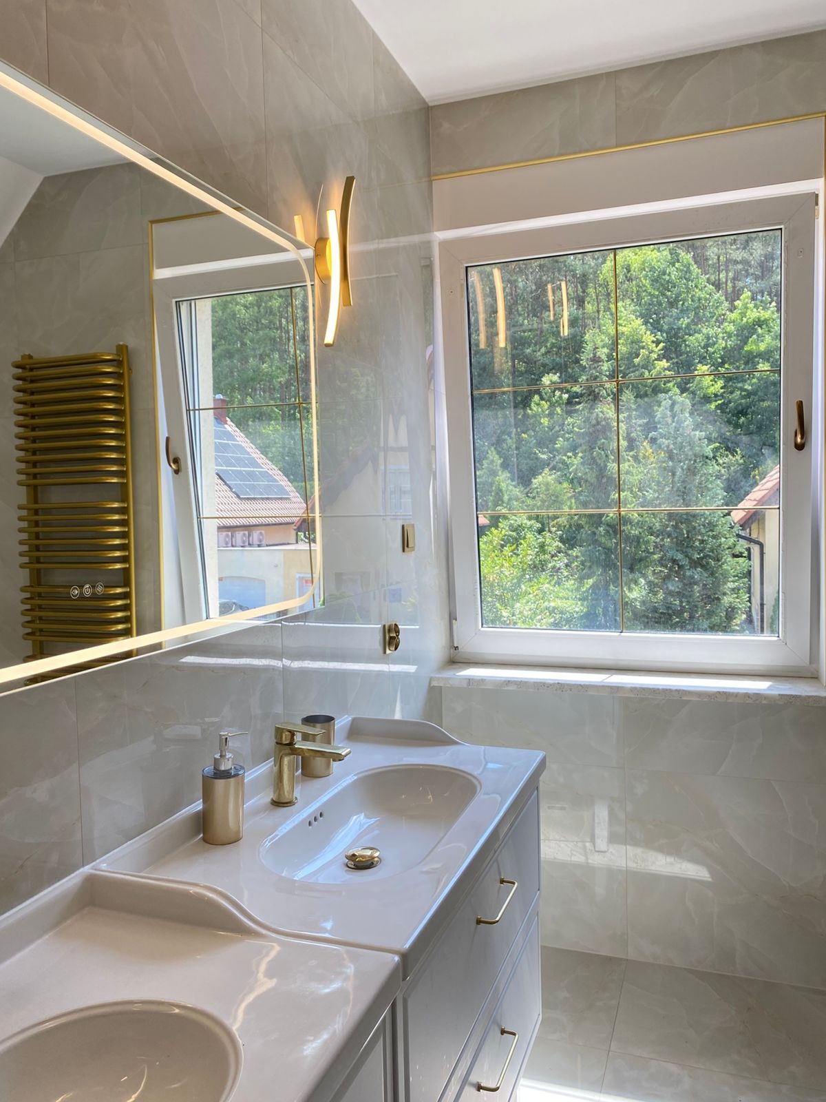 Jasna łazienka z marmurowymi płytkami, złotymi akcentami i dużym oknem z widokiem na zieleń. Umywalka z szafką, lustro z podświetleniem i grzejnik drabinkowy.