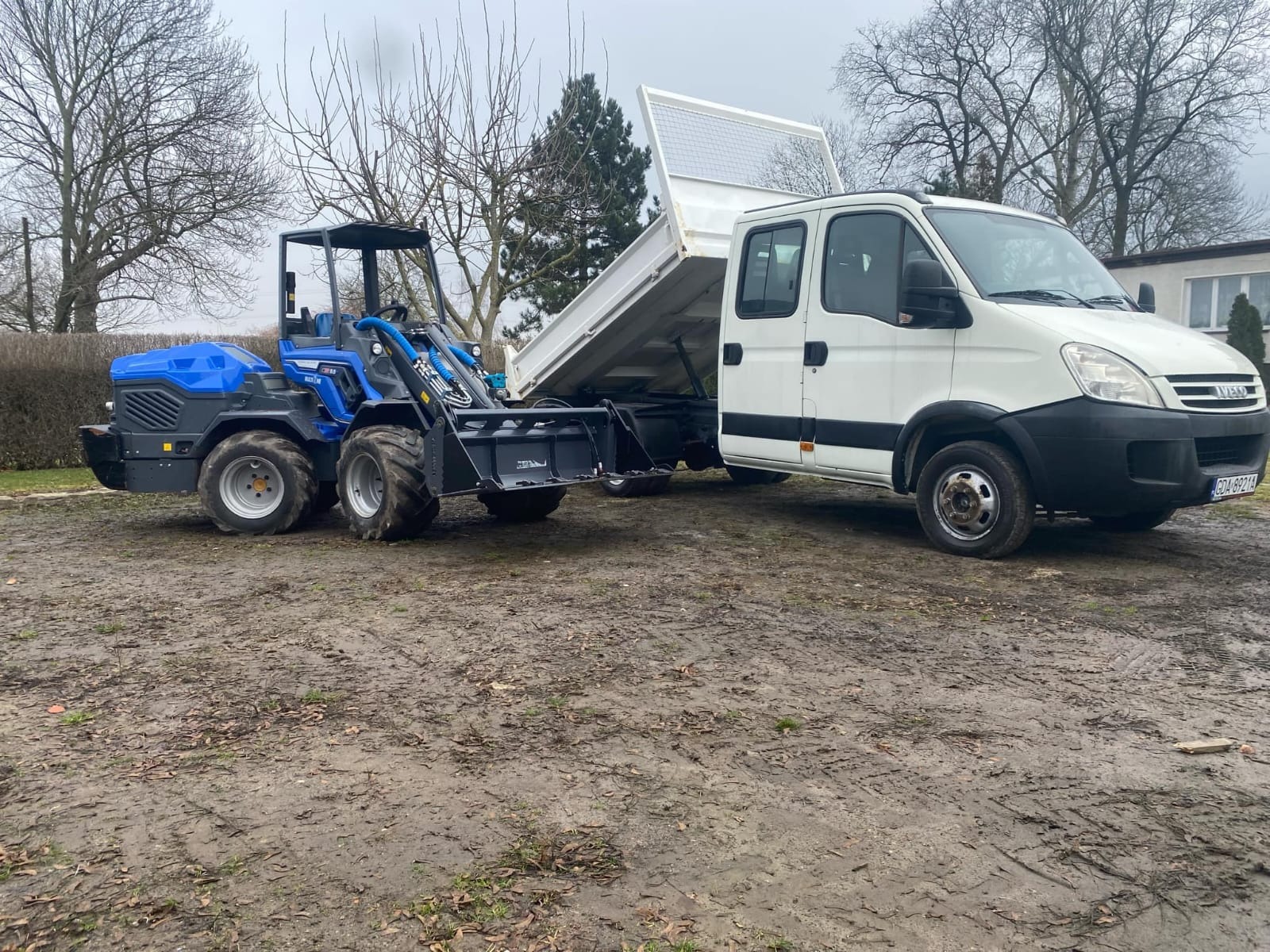 Niebieska miniładowarka przegubowa z łyżką i podniesiona skrzynia wywrotki białego samochodu ciężarowego na błotnistym terenie