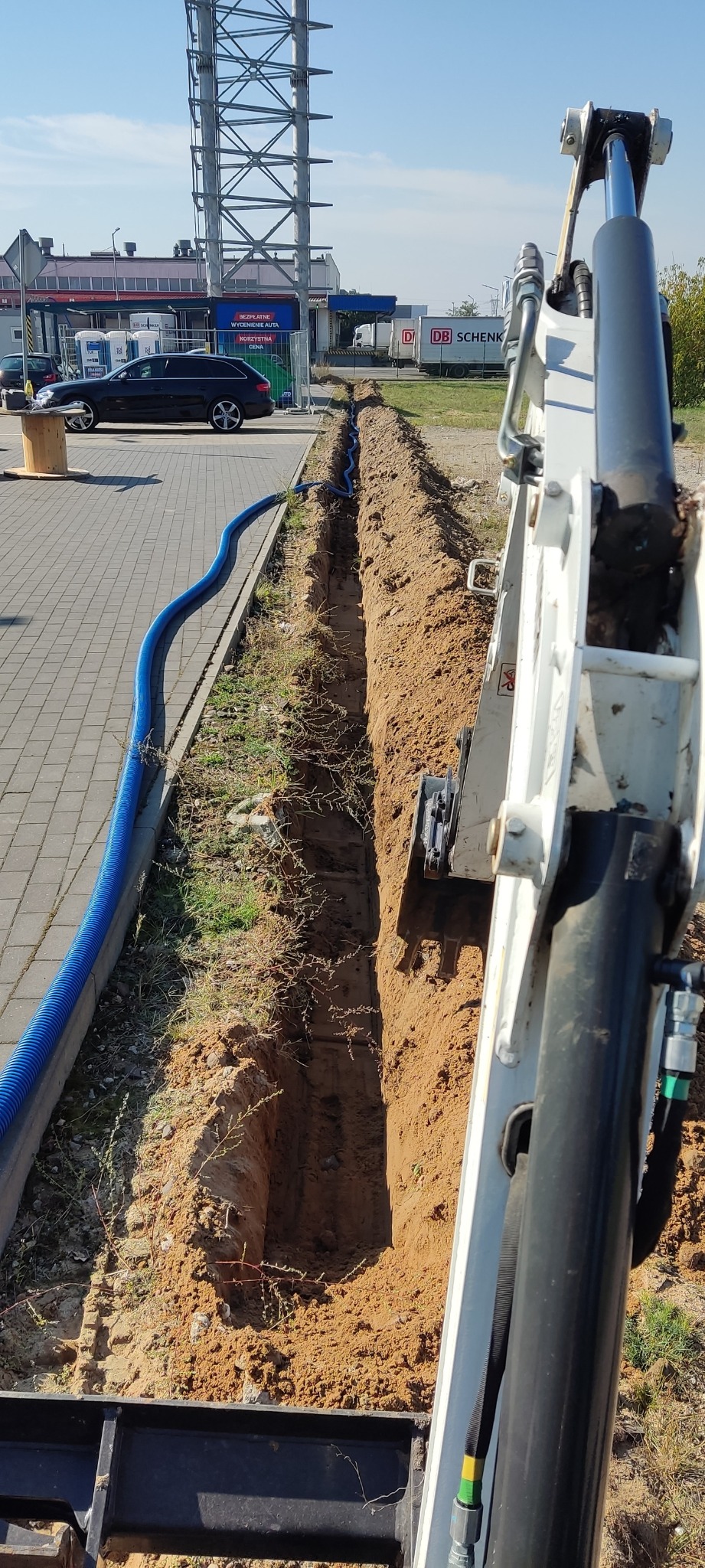 Wąski wykop w ziemi, z widoczną niebieską rurą karbowaną na dnie, wykonany minikoparką; w tle budynek z logo firmy transportowej i zaparkowane auto.