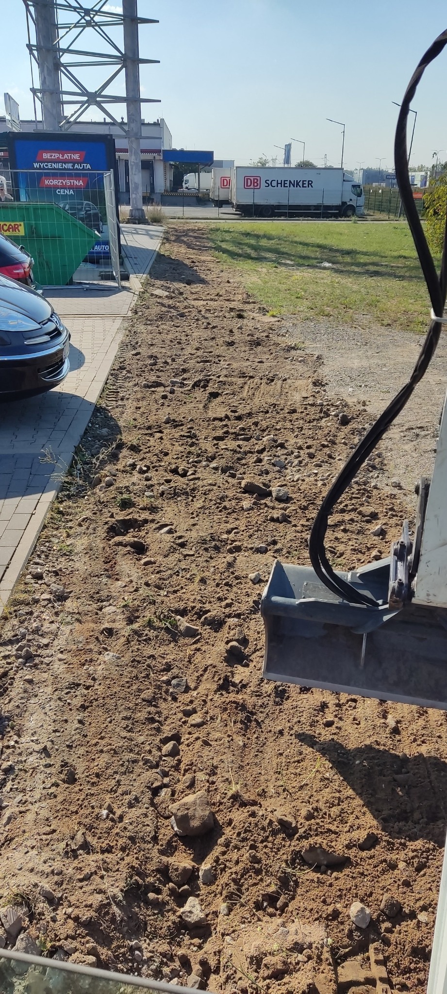 Równanie terenu przy użyciu łyżki koparki, widoczne nierówności gleby, kamienie i ślady po pracy maszyny, w tle budynki i zaparkowane samochody.