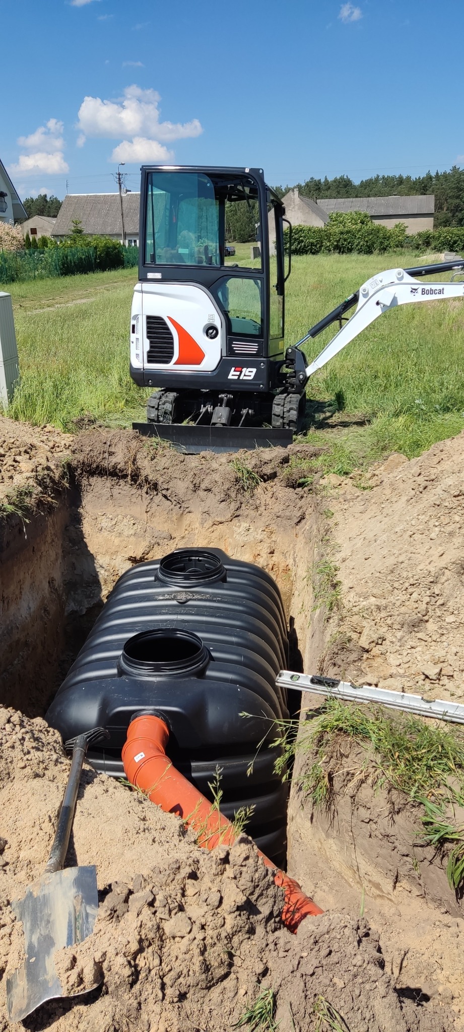 Czarny zbiornik bezodpływowy z pomarańczową rurą w wykopie, obok mini koparka Bobcat E19, poziomica i łopata, trawa i budynki w tle.