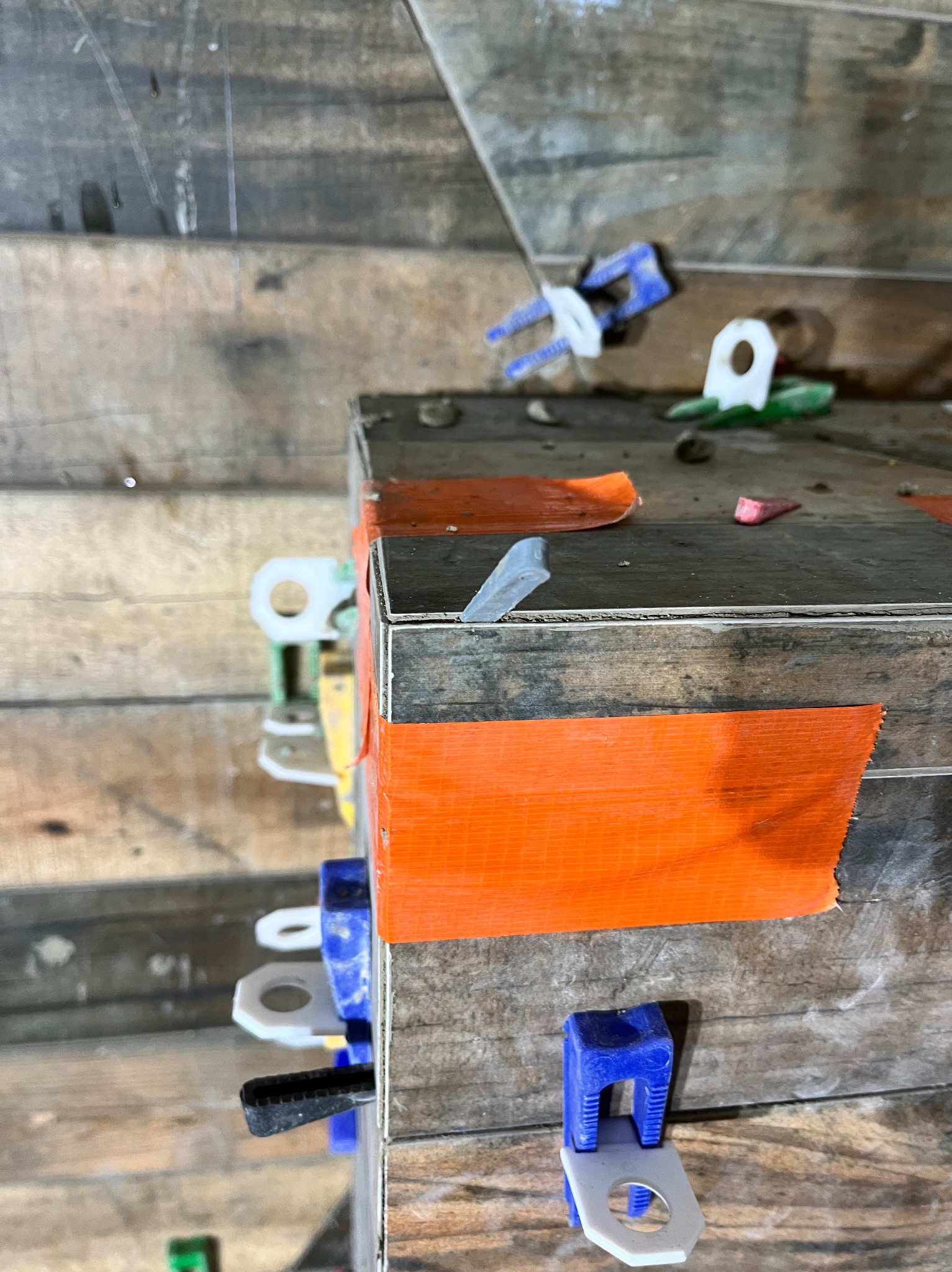 Fragment układanej powierzchni z płytek, widoczne klipsy poziomujące różnych kolorów i taśma maskująca, surowe drewniane tło.