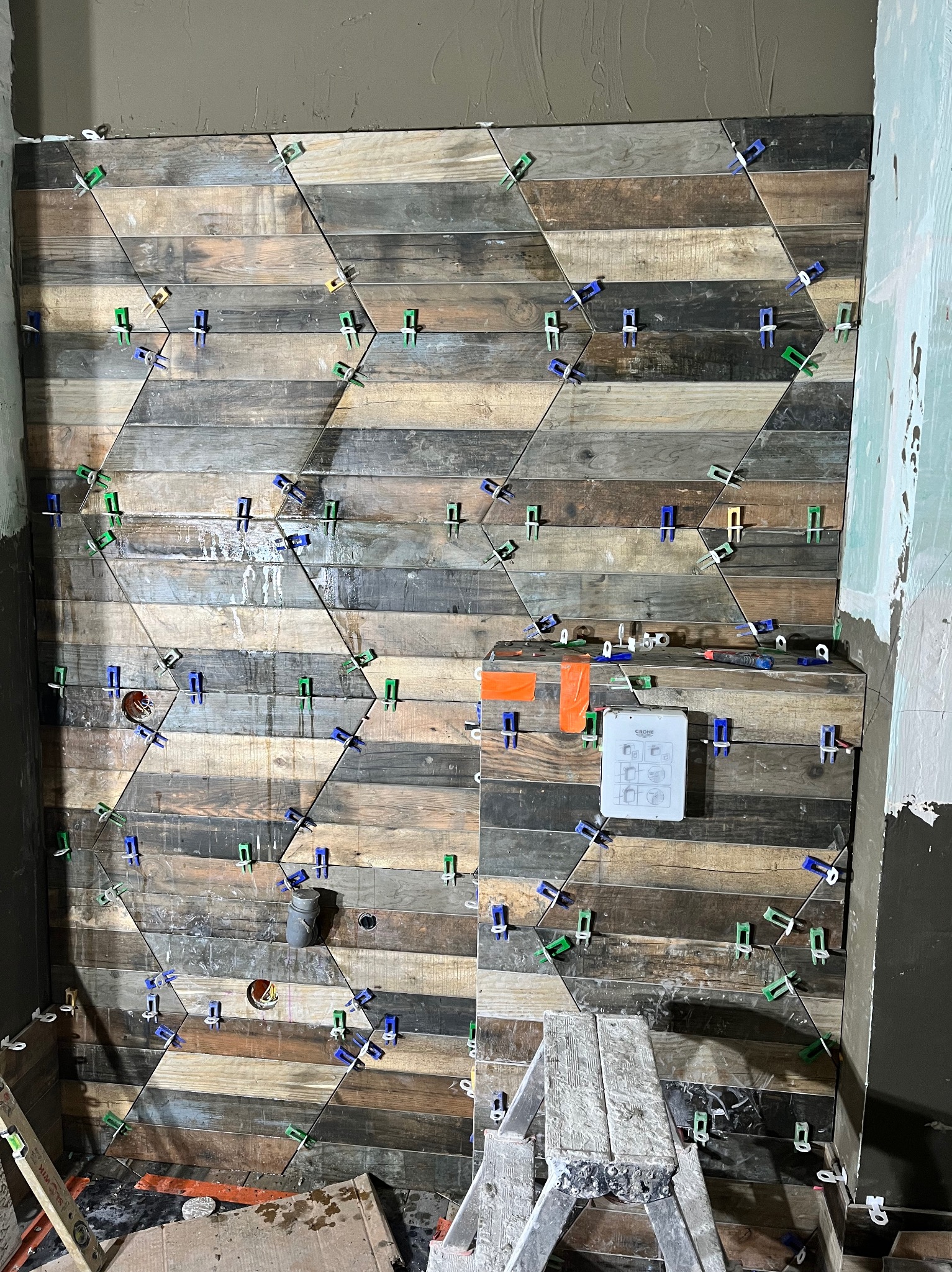 Ściana w trakcie układania płytek imitujących drewno w heksagonalny wzór, z użyciem klipsów poziomujących, widoczna instalacja wodno-kanalizacyjna i drabina malarska.