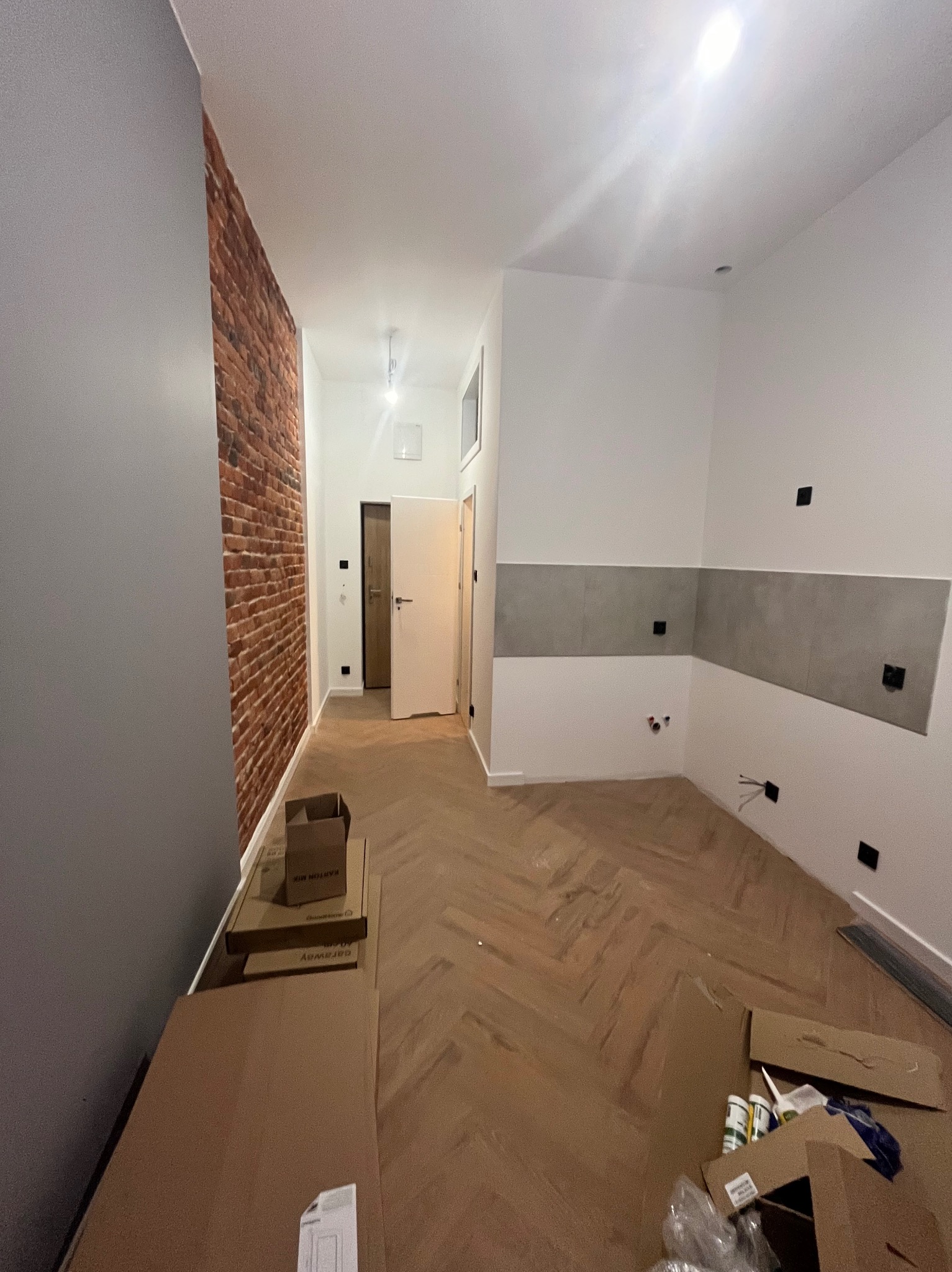 Wnętrze w trakcie prac wykończeniowych: ściana z cegły, świeżo ułożona podłoga w jodełkę, fragment ściany z szarymi płytkami, widoczne puszki elektryczne i materiały budowlane w kartonach.