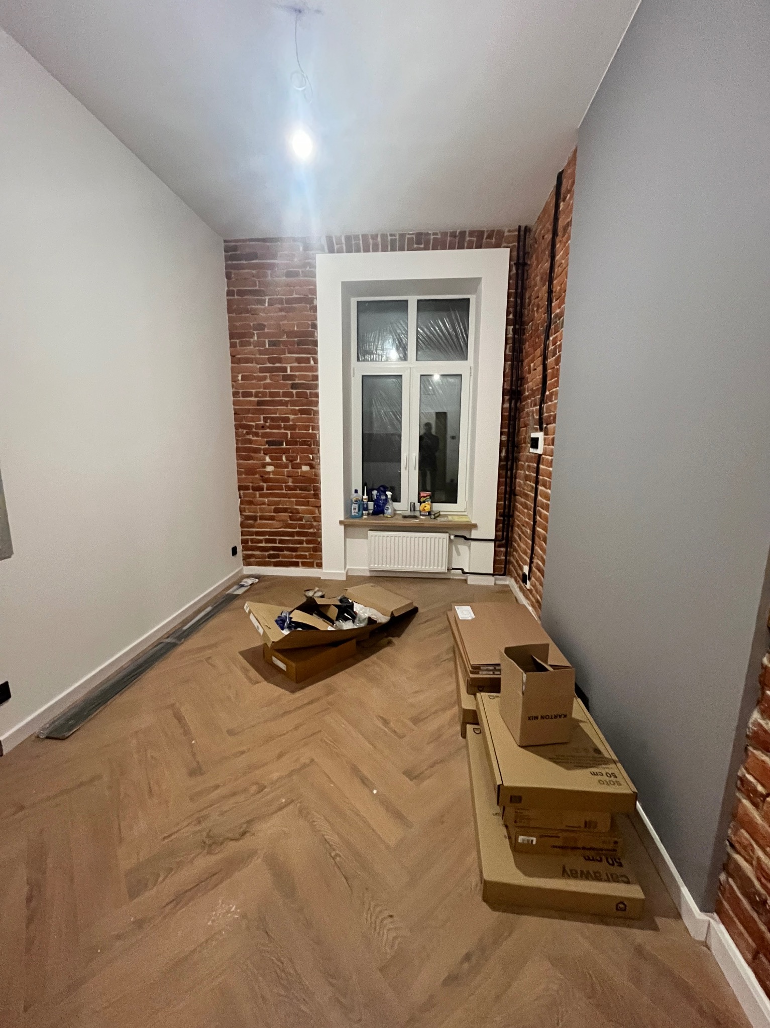Pokój w trakcie remontu z ułożonymi panelami podłogowymi w jodełkę, odsłoniętą cegłą przy oknie i na ścianie, kartonami na podłodze i przygotowanymi listwami przypodłogowymi.
