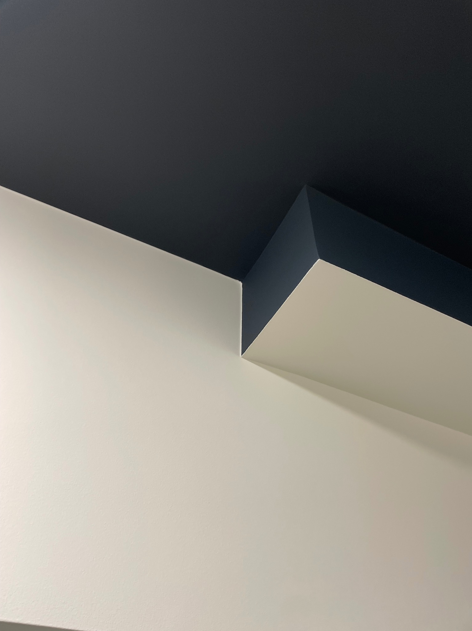 Pomieszczenie po malowaniu: kontrastowe połączenie białej ściany z ciemnogranatowym sufitem i wystającym fragmentem stropu, widoczne ostre krawędzie i gładkie powierzchnie.