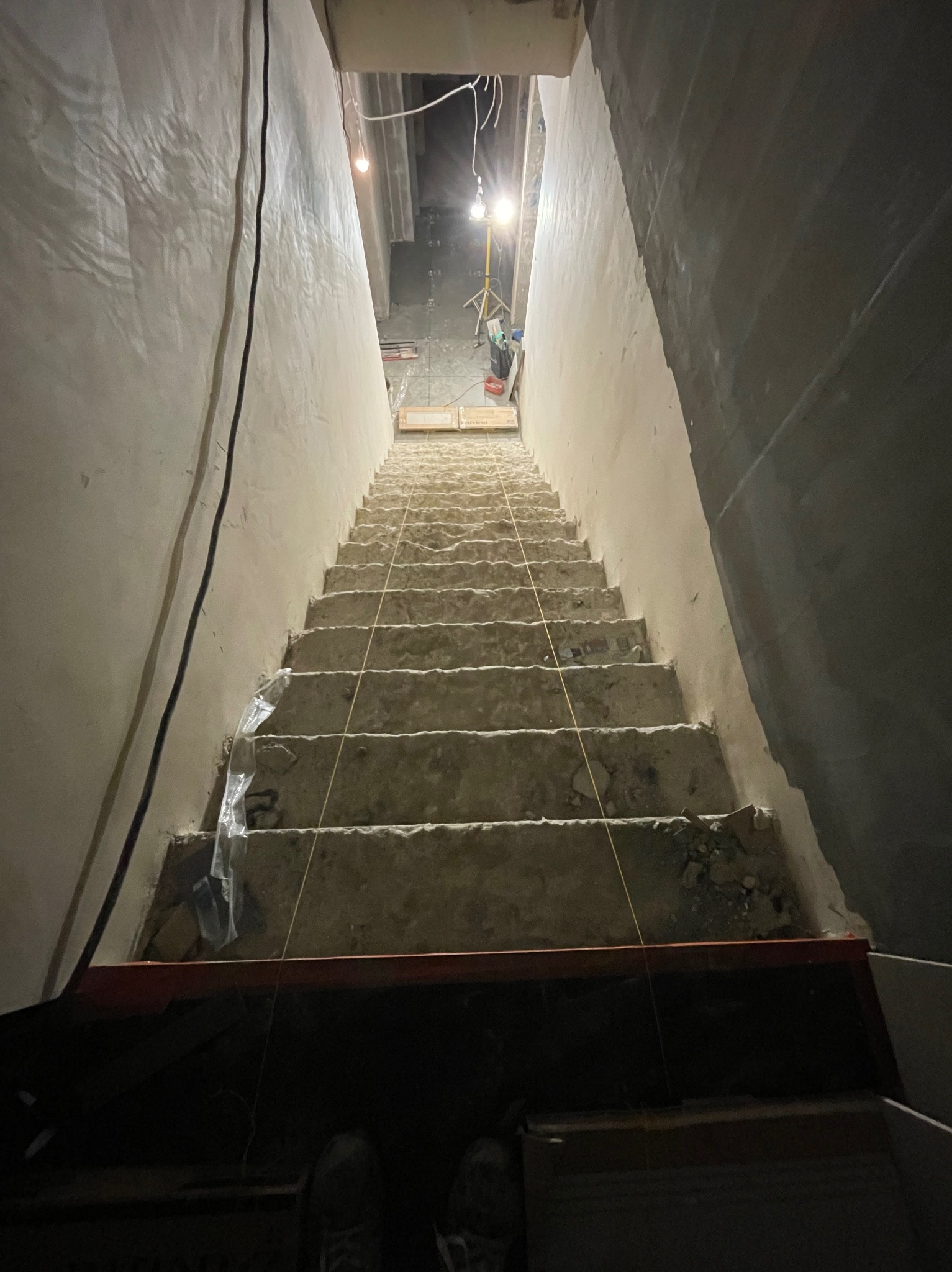 Schody w trakcie remontu, z widocznymi poziomymi liniami wyznaczającymi poziom, surowe ściany w różnej fazie wykończenia, oświetlenie budowlane w tle.