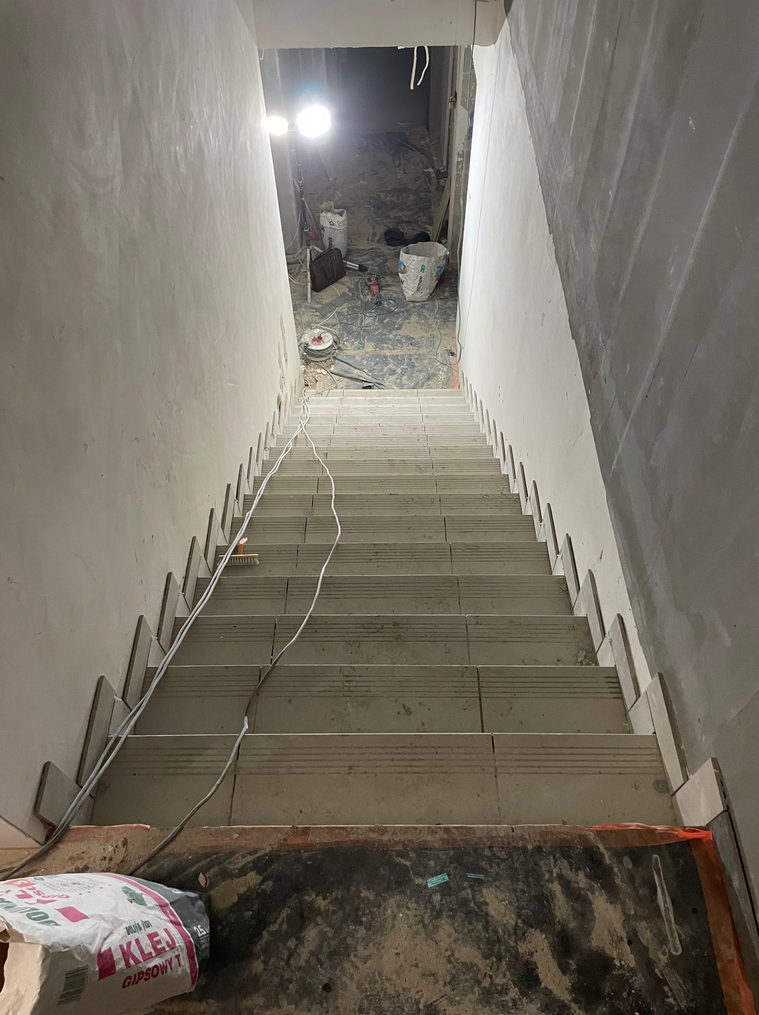 Schody w trakcie wykańczania, widoczne płytki schodowe z poziomymi rowkami, listwy dystansowe przy ścianie, kabel oraz worek z klejem gipsowym w dolnej części kadru, w tle narzędzia i lampa...