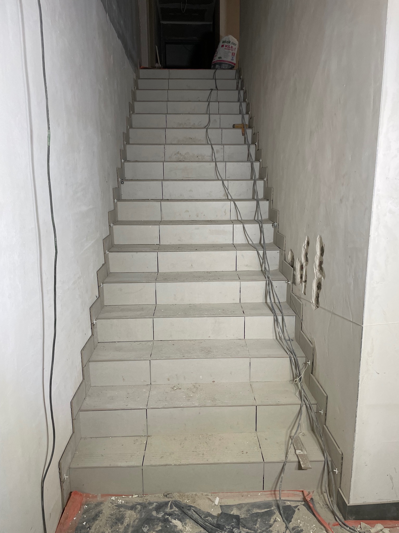 Klatka schodowa w trakcie układania płytek ceramicznych, widoczne krzyżyki dystansowe, przewody elektryczne i worek z klejem na górze schodów.