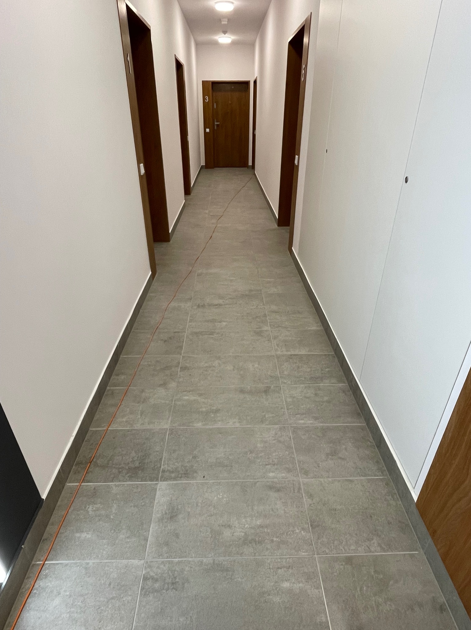 Wykończony korytarz w Łodzi z szarymi płytkami podłogowymi i białymi ścianami, widoczne drzwi do pokojów z numerami.