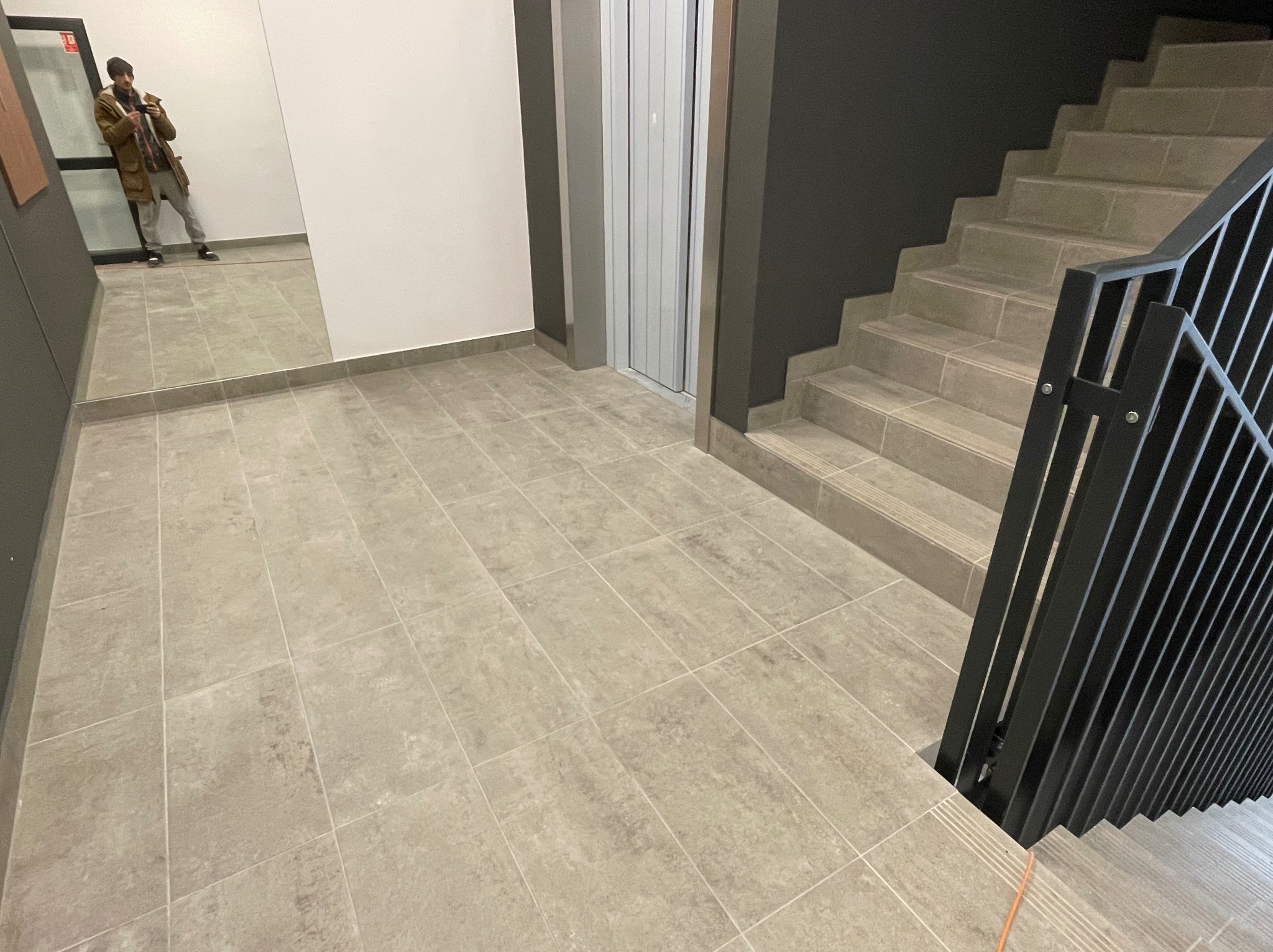 Beżowe płytki podłogowe w korytarzu budynku z windą i schodami, widoczny mężczyzna robiący zdjęcie w lustrze.