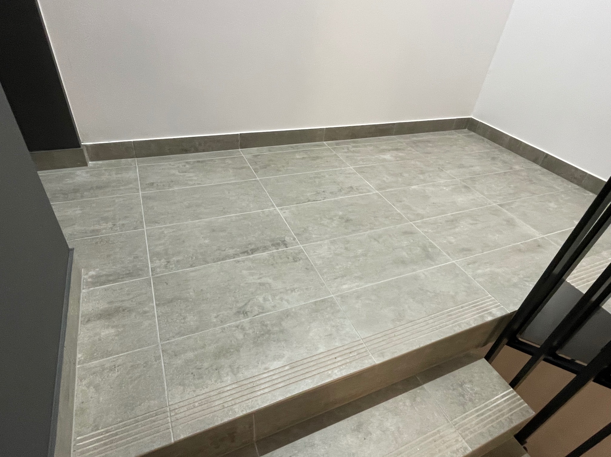 Szare, kwadratowe płytki podłogowe ułożone w korytarzu przy schodach z metalową barierką, widoczne listwy przypodłogowe.