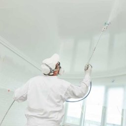 EXPERT REMONT SZCZECIN - Pracownik w kombinezonie ochronnym maluje sufit natryskowo w pomieszczeniu z listwami dekoracyjnymi przy suficie i oknami zabezpieczonymi folią.