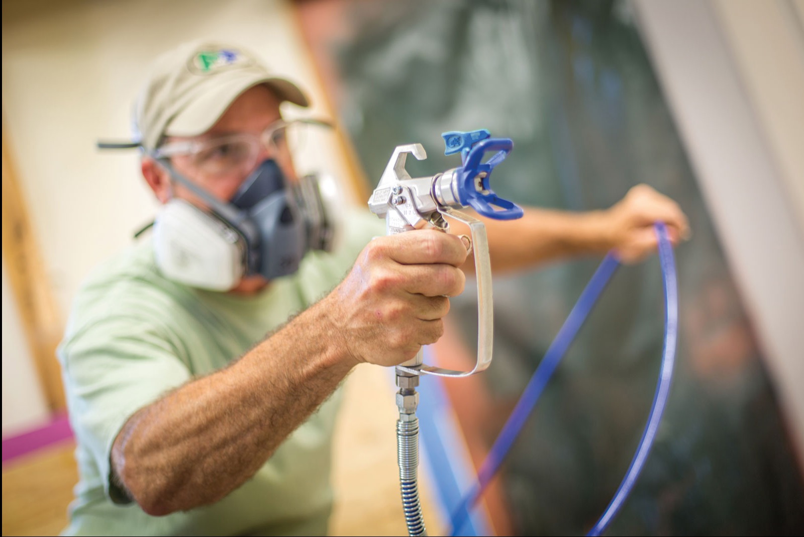Mężczyzna w masce i okularach ochronnych trzyma pistolet natryskowy z niebieskimi elementami, przygotowując się do malowania ściany.