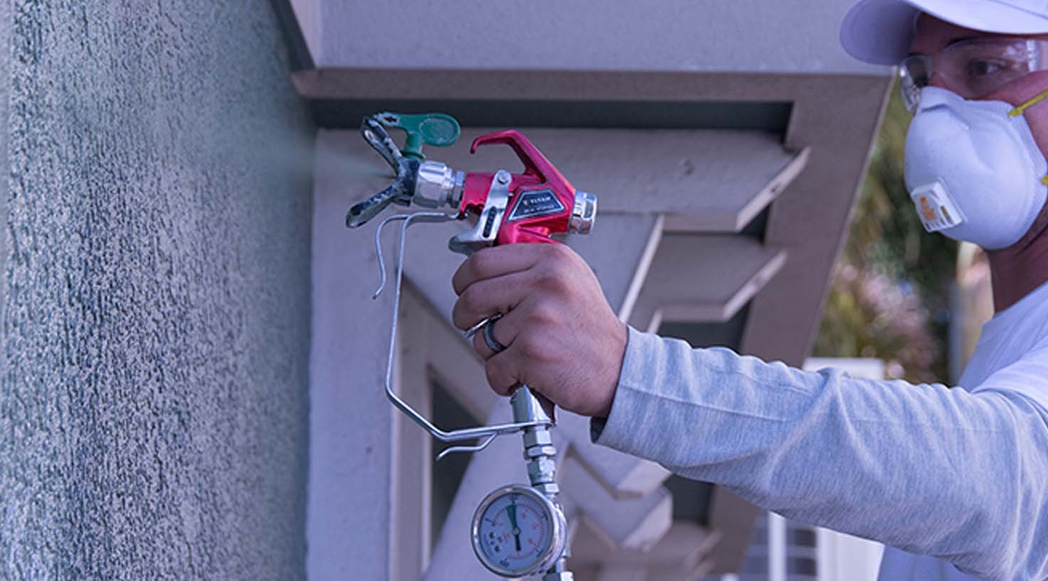 Pracownik w masce i okularach maluje ścianę natryskowo różowym pistoletem z manometrem.