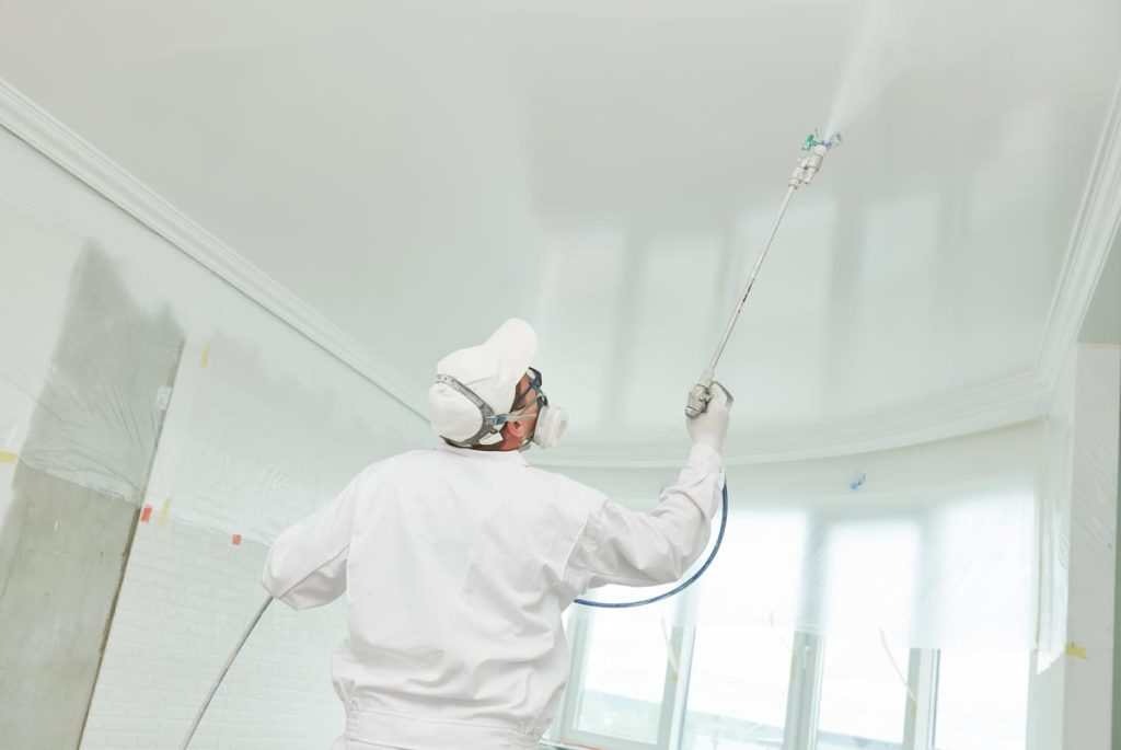 Pracownik w kombinezonie ochronnym maluje sufit natryskowo w pomieszczeniu z listwami dekoracyjnymi przy suficie i oknami zabezpieczonymi folią.
