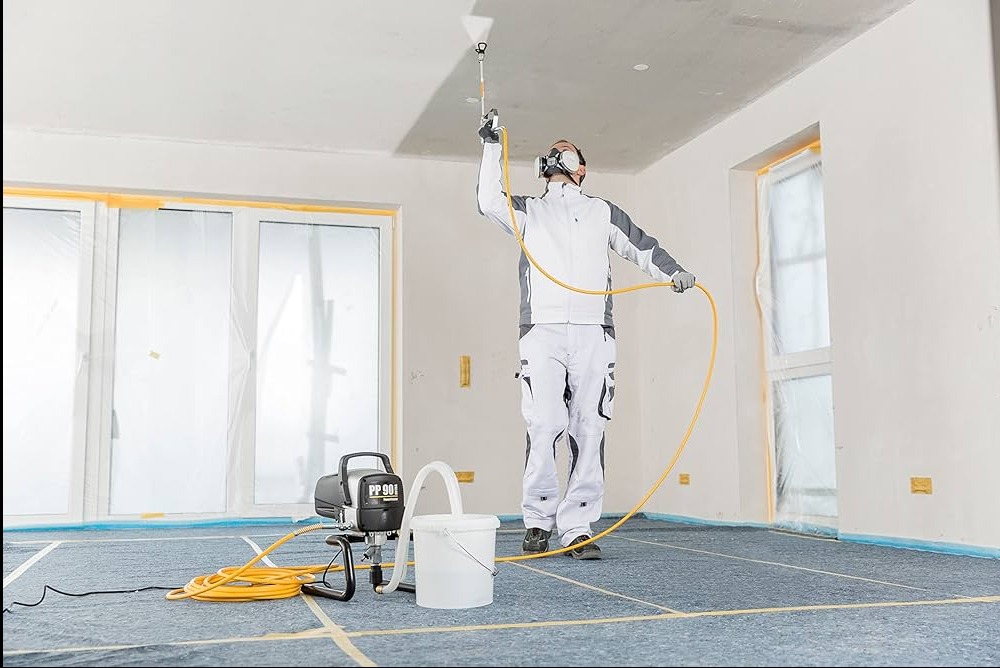 Pracownik w kombinezonie ochronnym maluje sufit agregatem natryskowym PP 90 i maską, podłoga zabezpieczona folią malarską, okna zakryte folią ochronną.