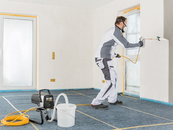 Pracownik w masce maluje ścianę natryskowo, używając agregatu malarskiego PP 90 i zabezpieczając okna folią oraz podłogę kartonem.