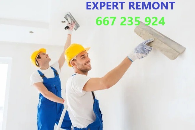 Dwóch uśmiechniętych pracowników w niebieskich kombinezonach i żółtych kaskach nakłada gładź na ścianę za pomocą packi, jeden stoi na drabinie.