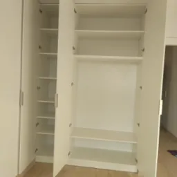 Biała, otwarta szafa wnękowa z p&oacute;łkami i drążkiem na ubrania, widok z przodu, jasne wnętrze, parkiet na podłodze. Minimalistyczny design.