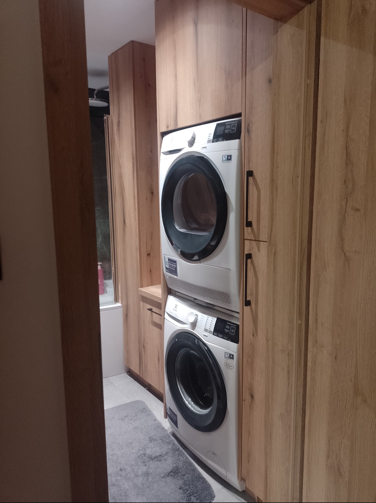 Pralka i suszarka w zabudowie z drewnopodobnych paneli, widoczne z boku w łazience z szarym dywanikiem i prysznicem w tle.