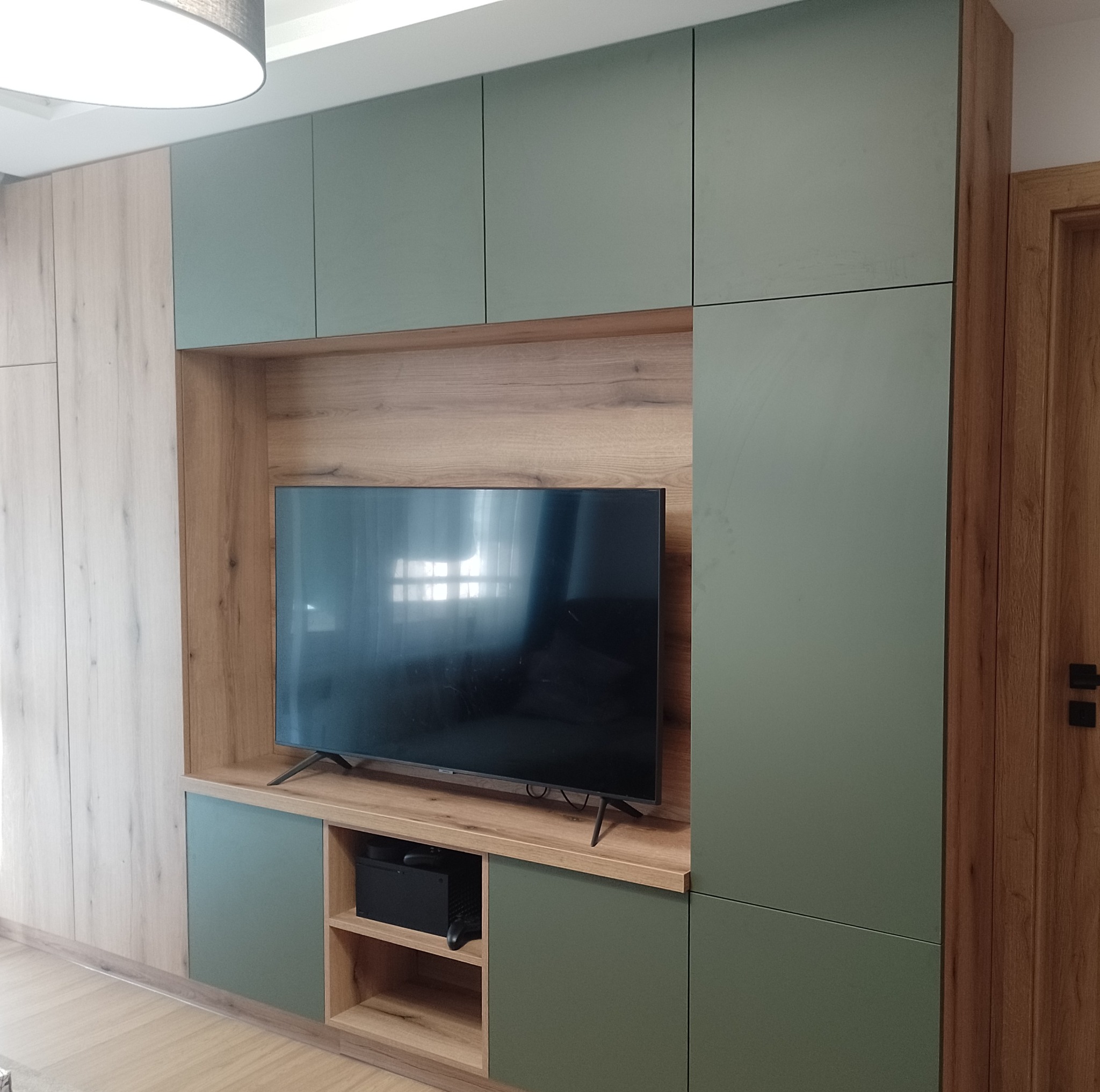 Nowoczesna szafa wnękowa z zielonymi frontami i drewnianym wykończeniem, wbudowana przestrzeń na telewizor, widoczna konsola do gier.