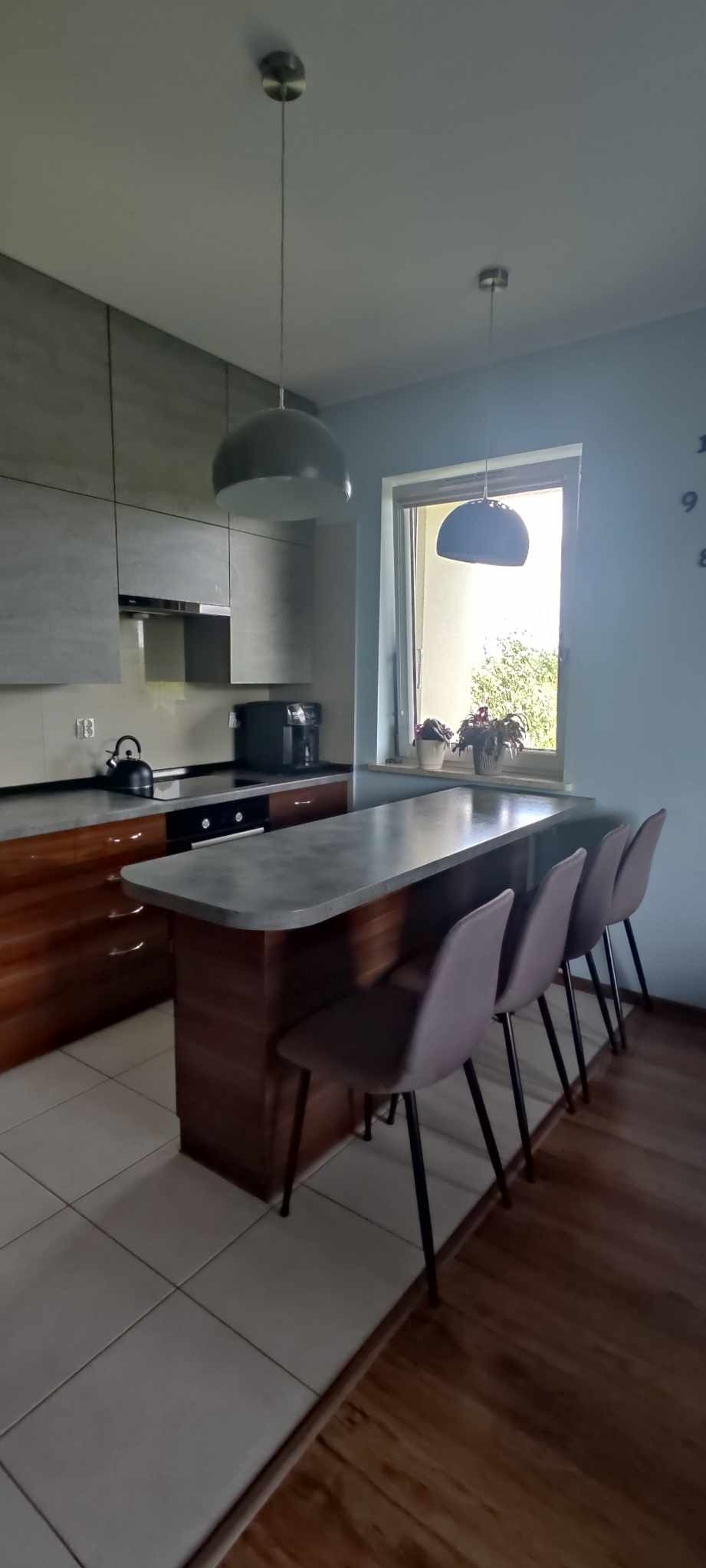 Nowoczesna kuchnia z wyspą i czterema hokerami w odcieniach szarości i brązu, oświetlona wiszącymi lampami z widokiem na zieleń za oknem.