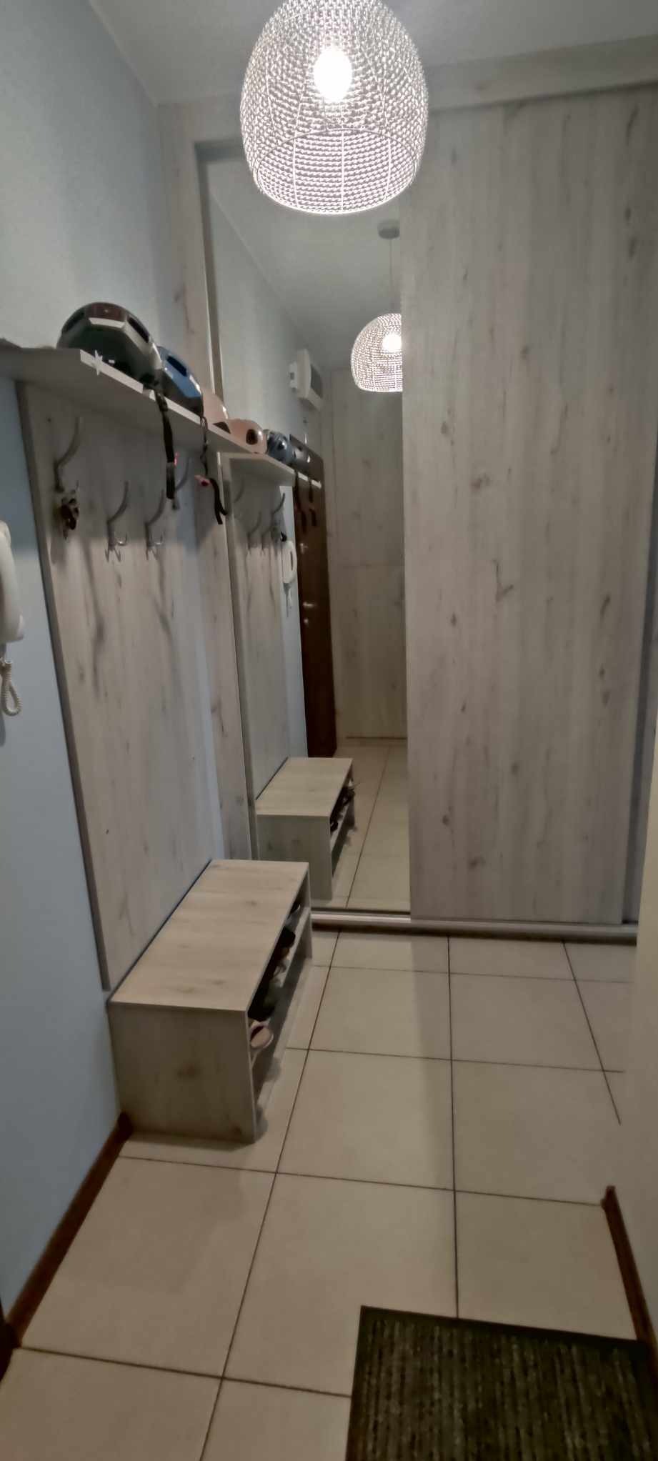 Wnętrze przedpokoju z szafą przesuwną z lustrem, wieszakiem z półką na kaski rowerowe i szafką na buty z siedziskiem, oświetlone ażurową lampą wiszącą.