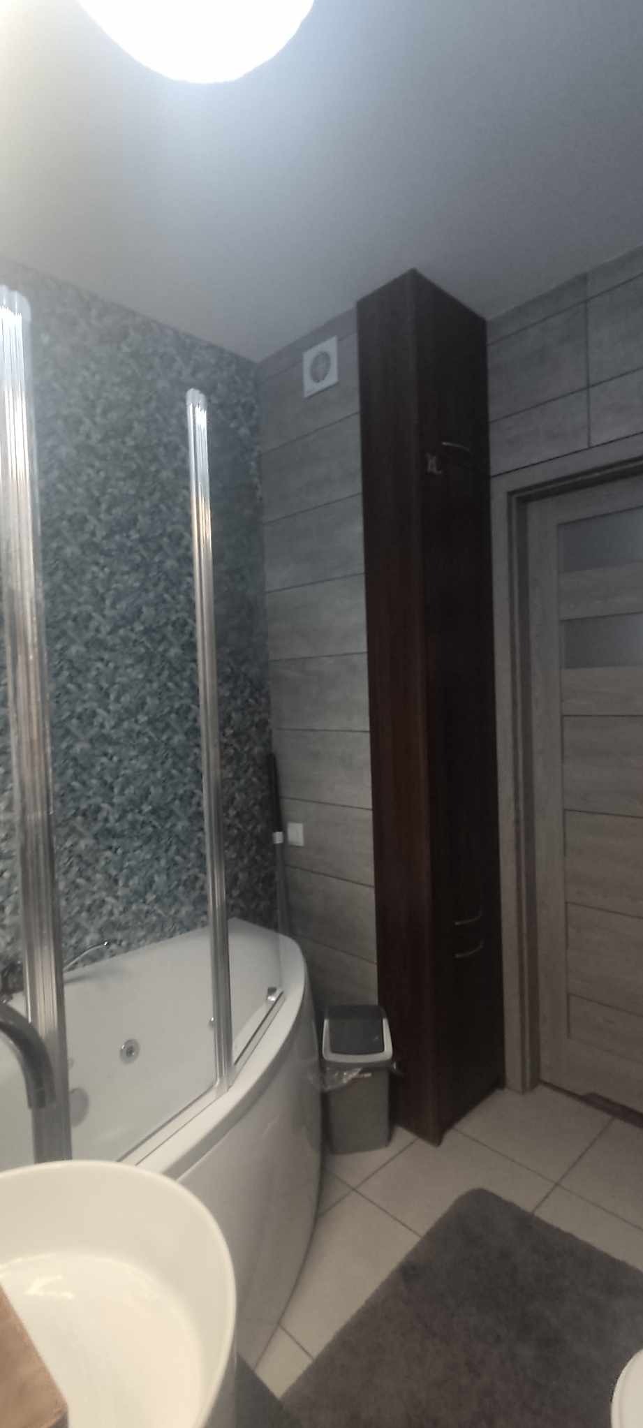 Nowoczesna łazienka z narożną wanną z hydromasażem za przeszkloną kabiną, wysoka, ciemna szafka łazienkowa i szare płytki na ścianach.