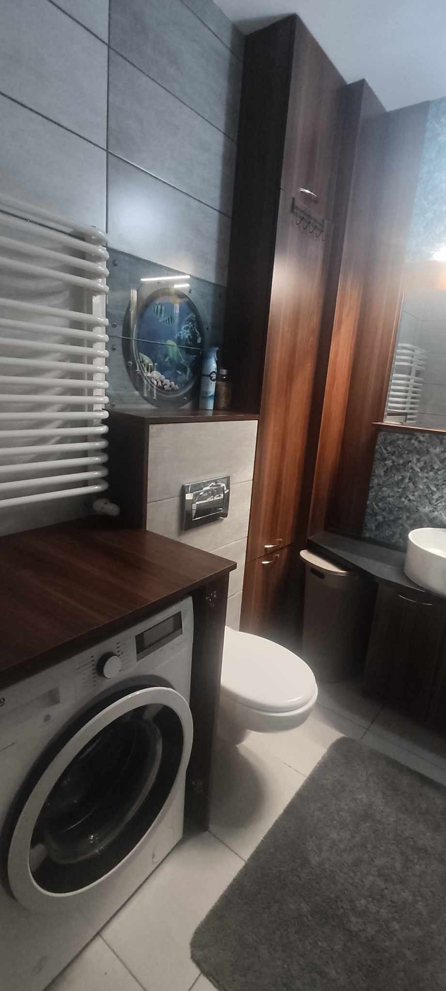 Nowoczesna łazienka z pralką pod blatem z ciemnego drewna, grzejnikiem drabinkowym, toaletą i szafką z drewnianymi frontami oraz okrągłym elementem dekoracyjnym z motywem akwarium.