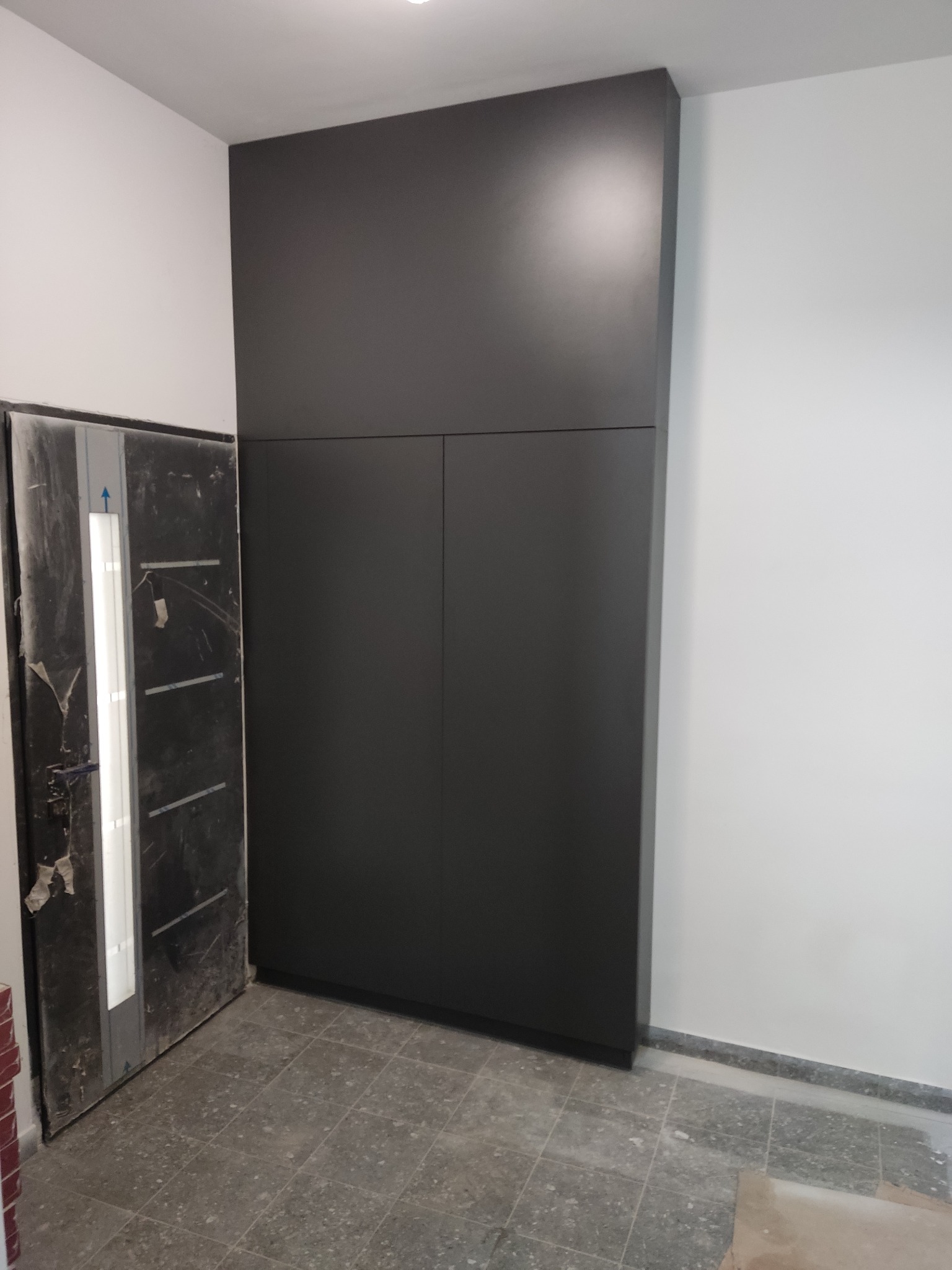 Czarna szafa wnękowa z dwiema parami drzwi, zamontowana w przedpokoju, widoczna obok brudnych drzwi wejściowych w trakcie remontu.