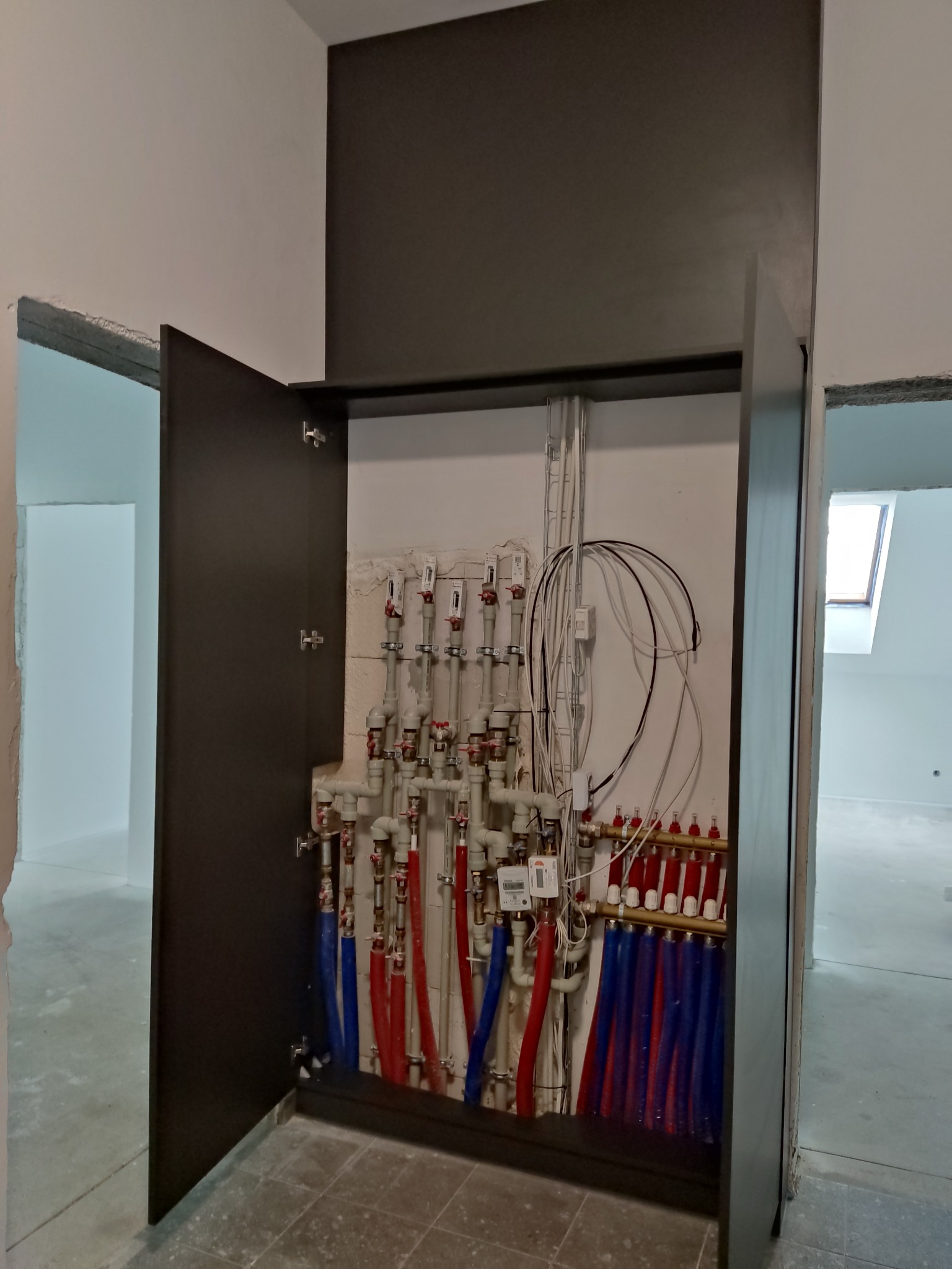 Rozdzielacz ogrzewania podłogowego ukryty w szafce wnękowej z otwartymi drzwiami, widoczne czerwone i niebieskie rury oraz liczniki, surowy stan wykończenia wnętrza.