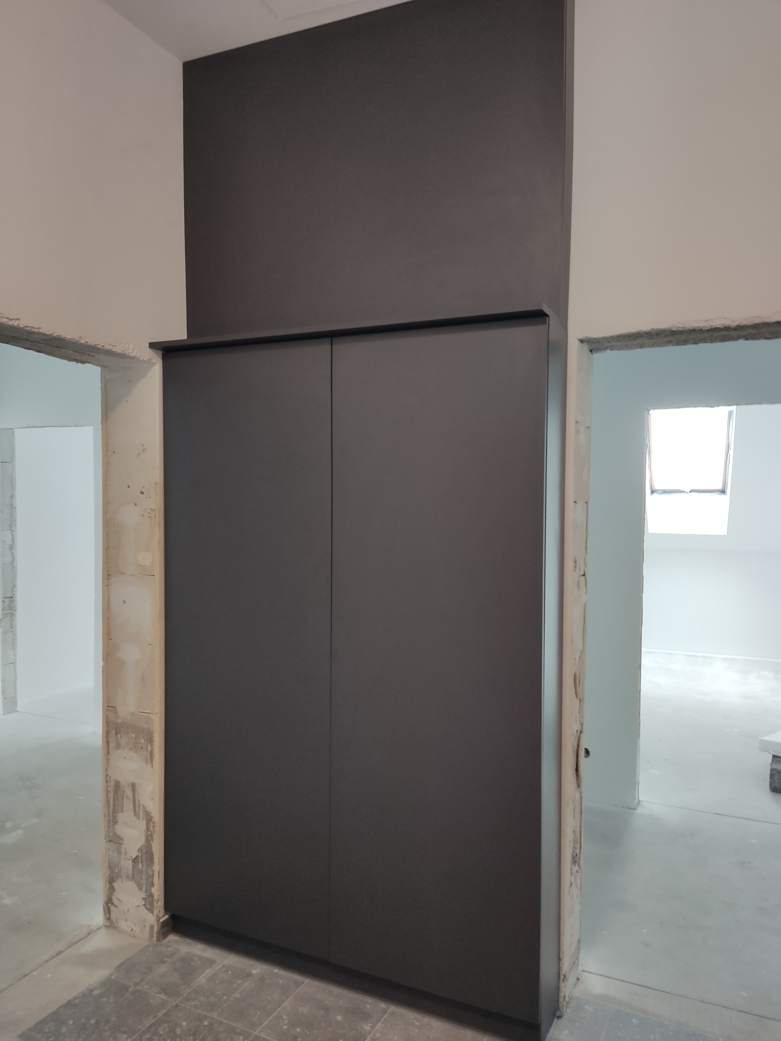 Ciemnoszara szafa wnękowa z dwoma drzwiami, zamontowana w surowym stanie wykończenia wnętrza, widoczne fragmenty niezabezpieczonych ścian i podłogi.