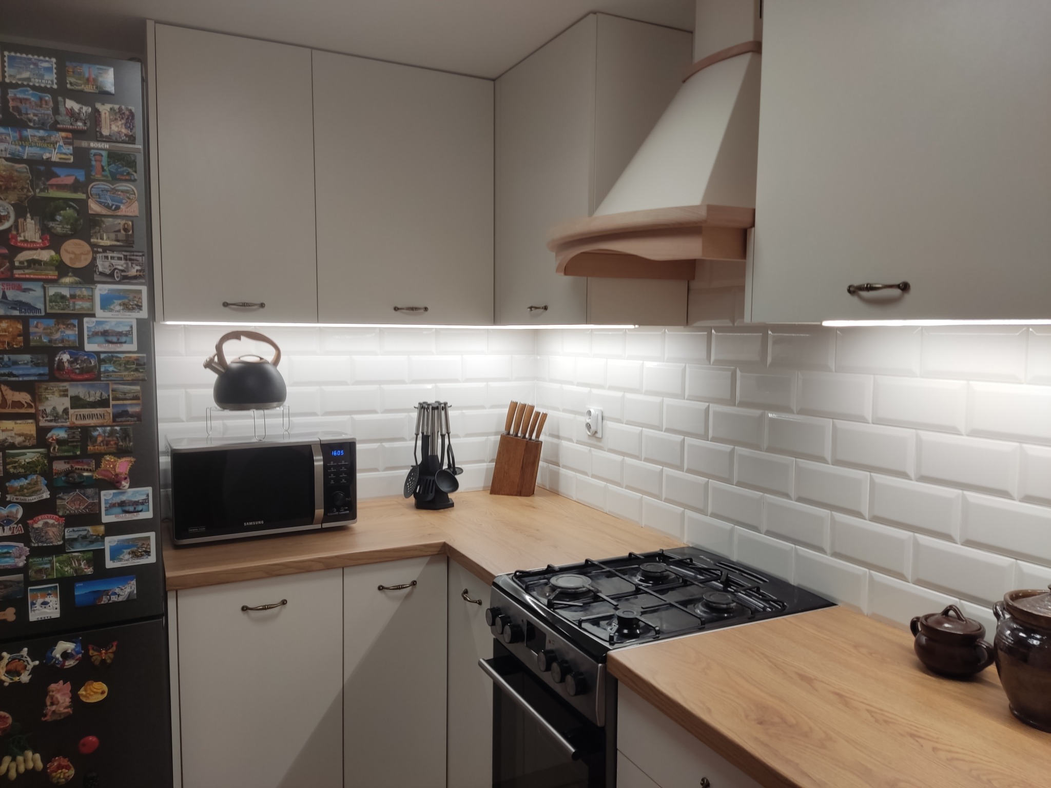 Jasna kuchnia z drewnianym blatem i białymi płytkami metro. Lodówka z magnesami, kuchenka gazowa i drewniane akcenty na okapie dodają ciepła.