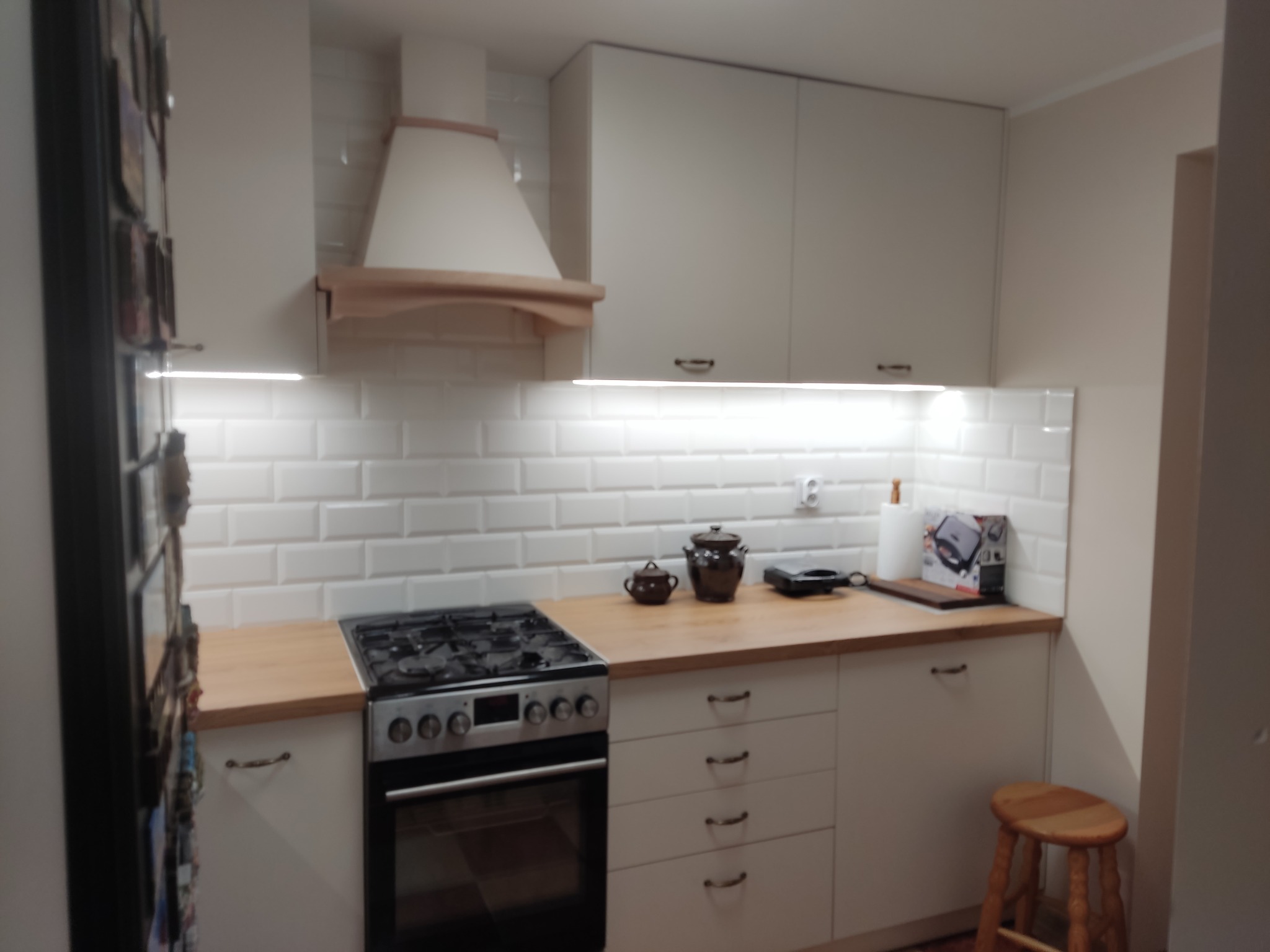 Jasna kuchnia na wymiar z białymi szafkami, drewnianym blatem i okapem z elementami drewna. Na blacie stoją garnki i inne akcesoria kuchenne.