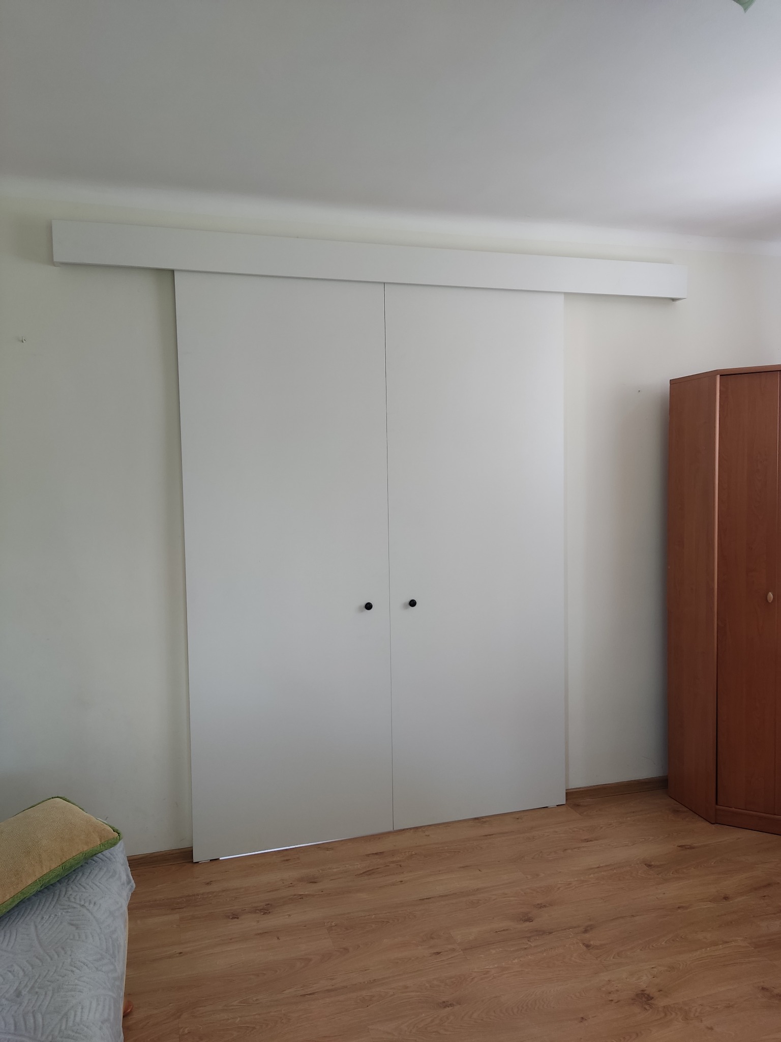 Minimalistyczne, białe drzwi przesuwne z czarnymi gałkami na tle jasnej ściany i drewnianej podłogi. Obok widoczny fragment szafy.